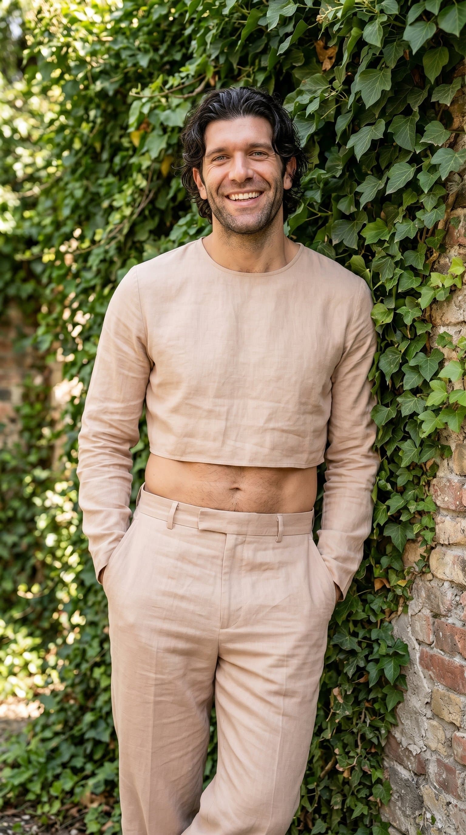 man in a nude high-waisted trouser and cropped top ensemble, standing against an ivy-covered wall, smiling brightly