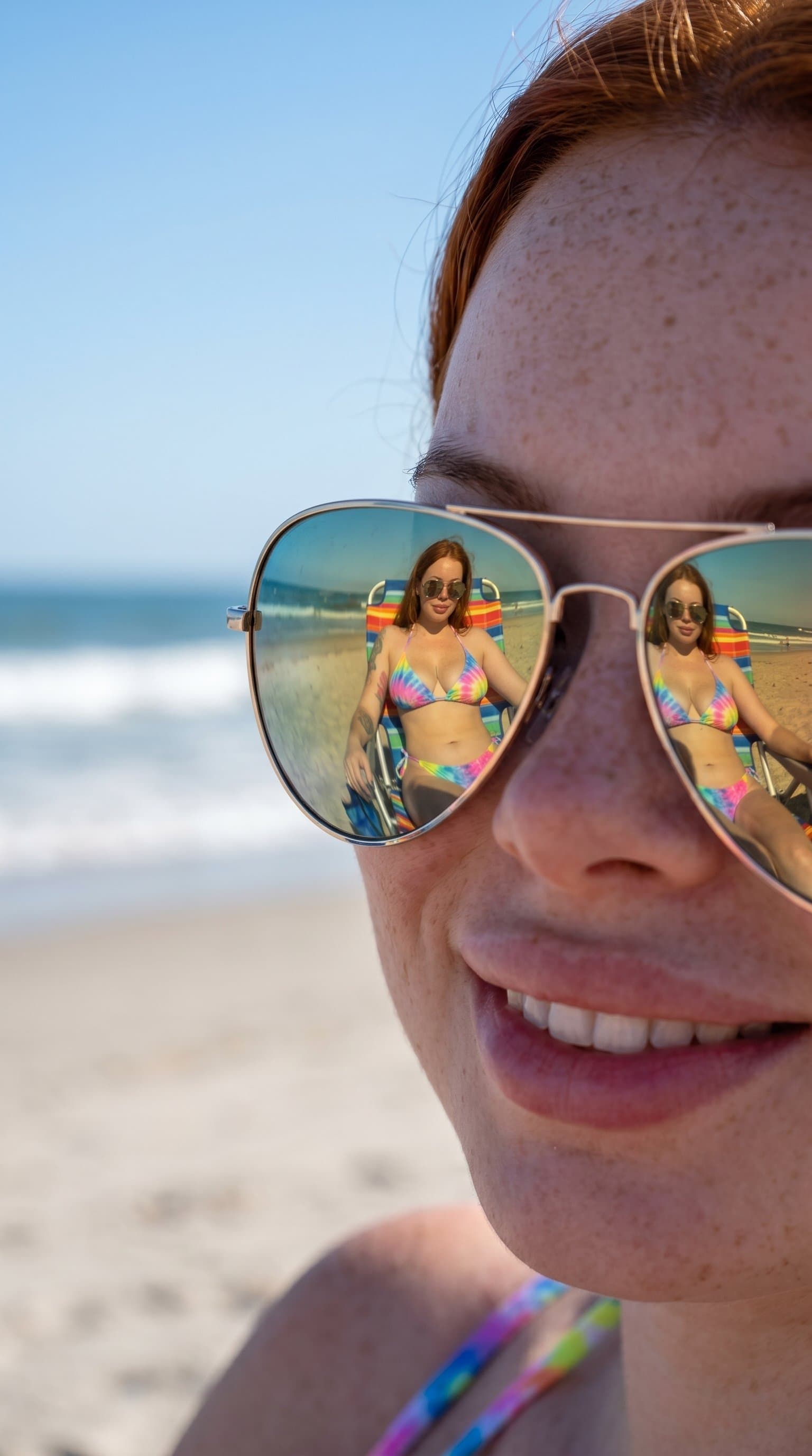 A woman in a tie-dye bikini, sitting on a beach chair, with their reflection mirrored on a pair of aviator sunglasses