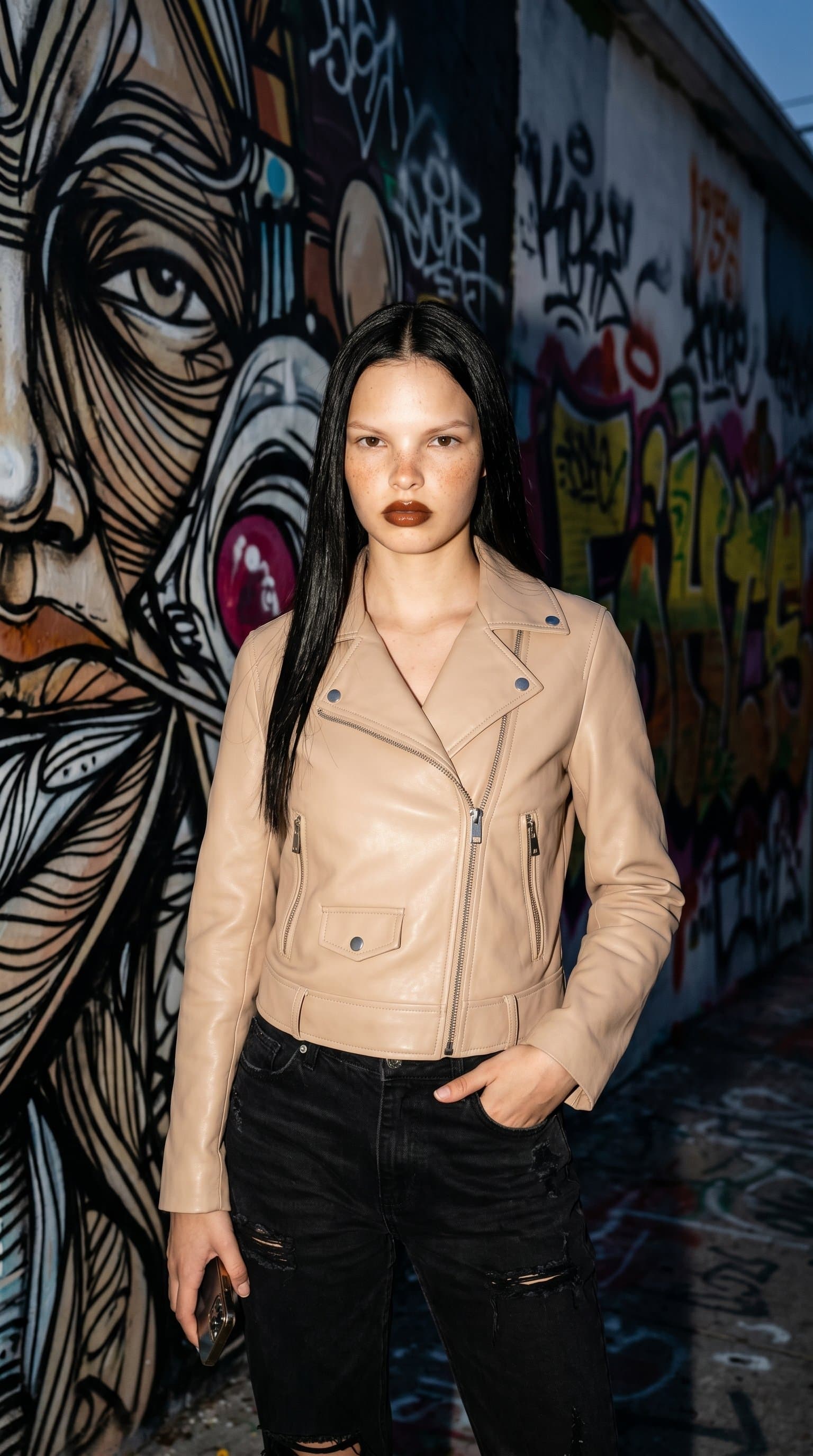 woman in a nude leather jacket and jeans posing against a graffiti wall, with dynamic, high contrast lighting emphasizing edginess