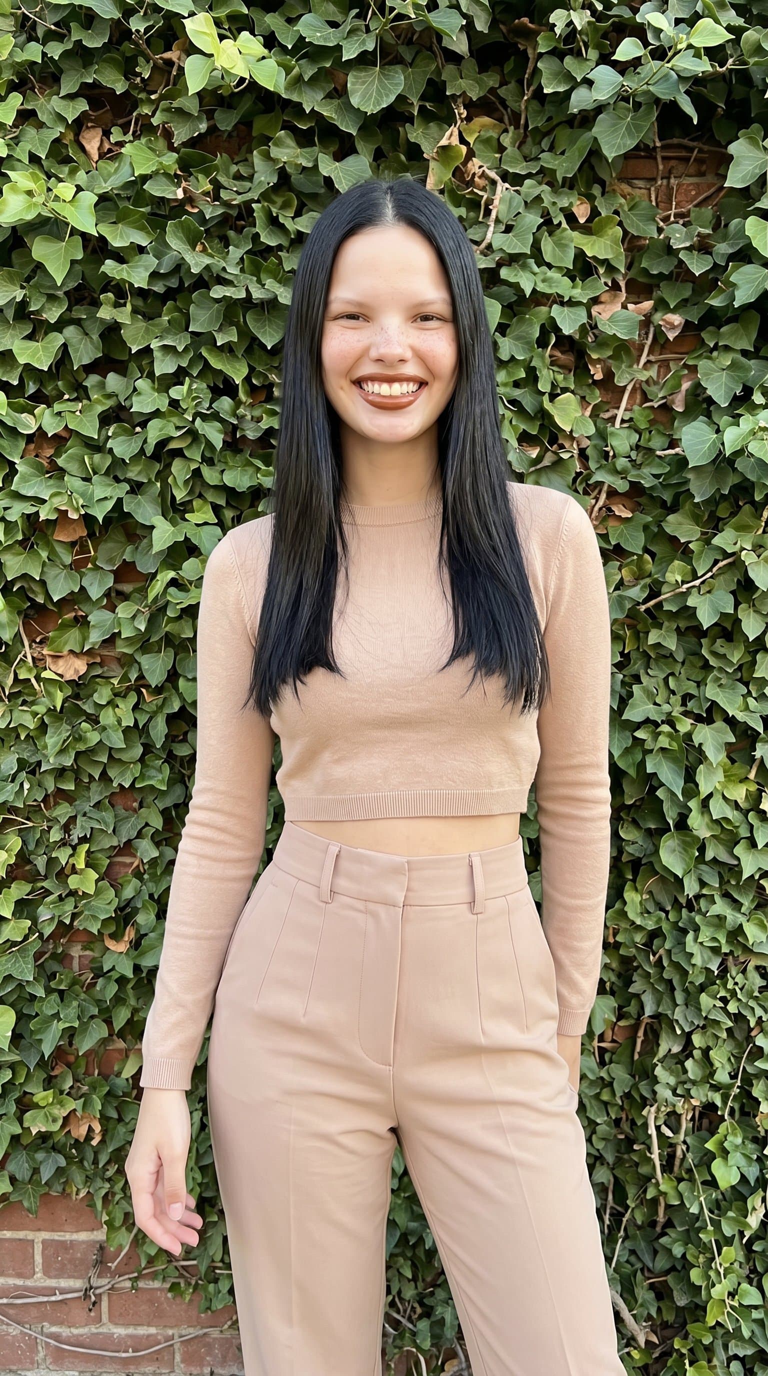 woman in a nude high-waisted trouser and cropped top ensemble, standing against an ivy-covered wall, smiling brightly