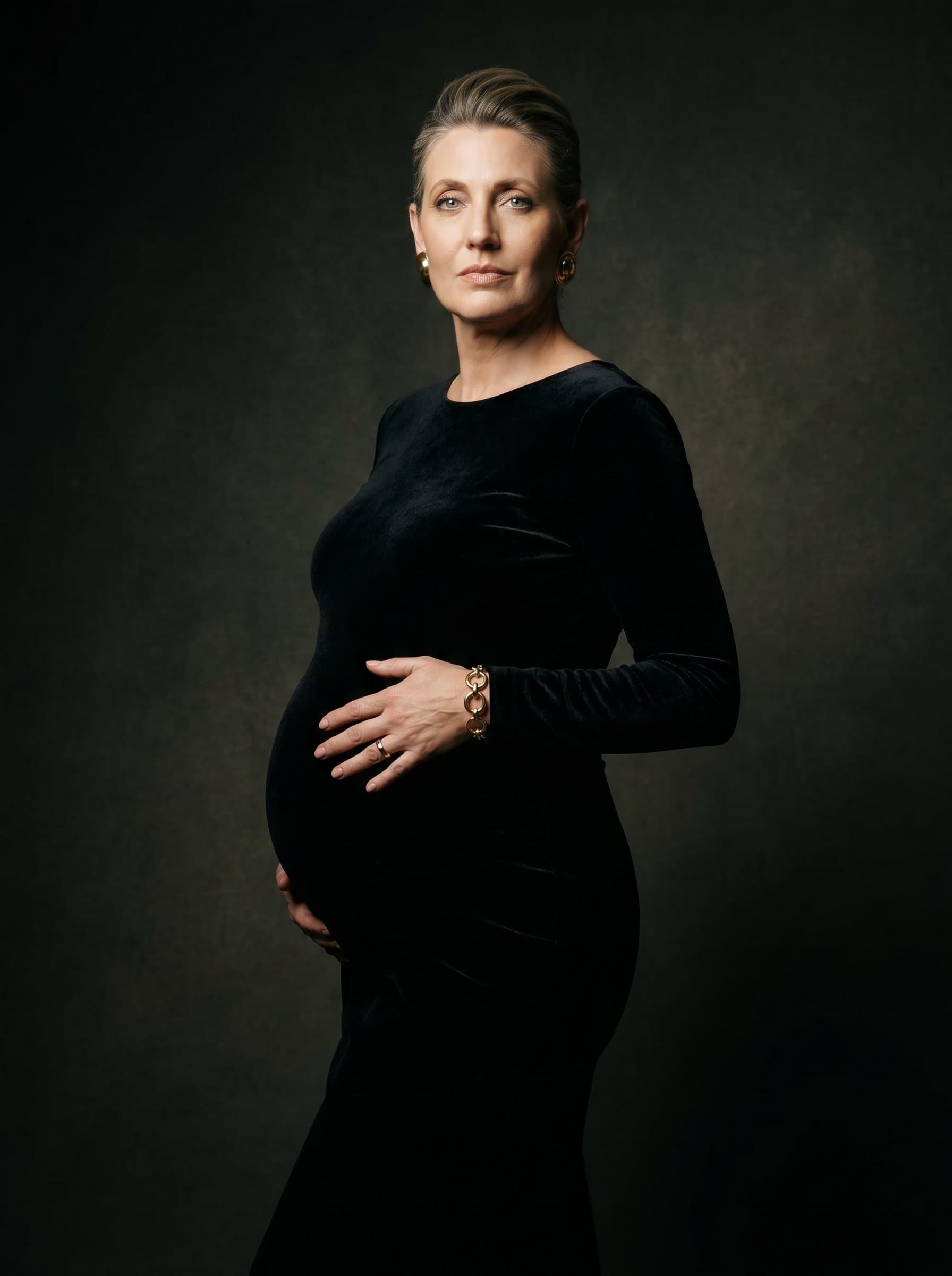 Pregnant in elegant black dress, dramatic studio lighting, one hand on belly, confident and radiant expression, high fashion maternity portrait