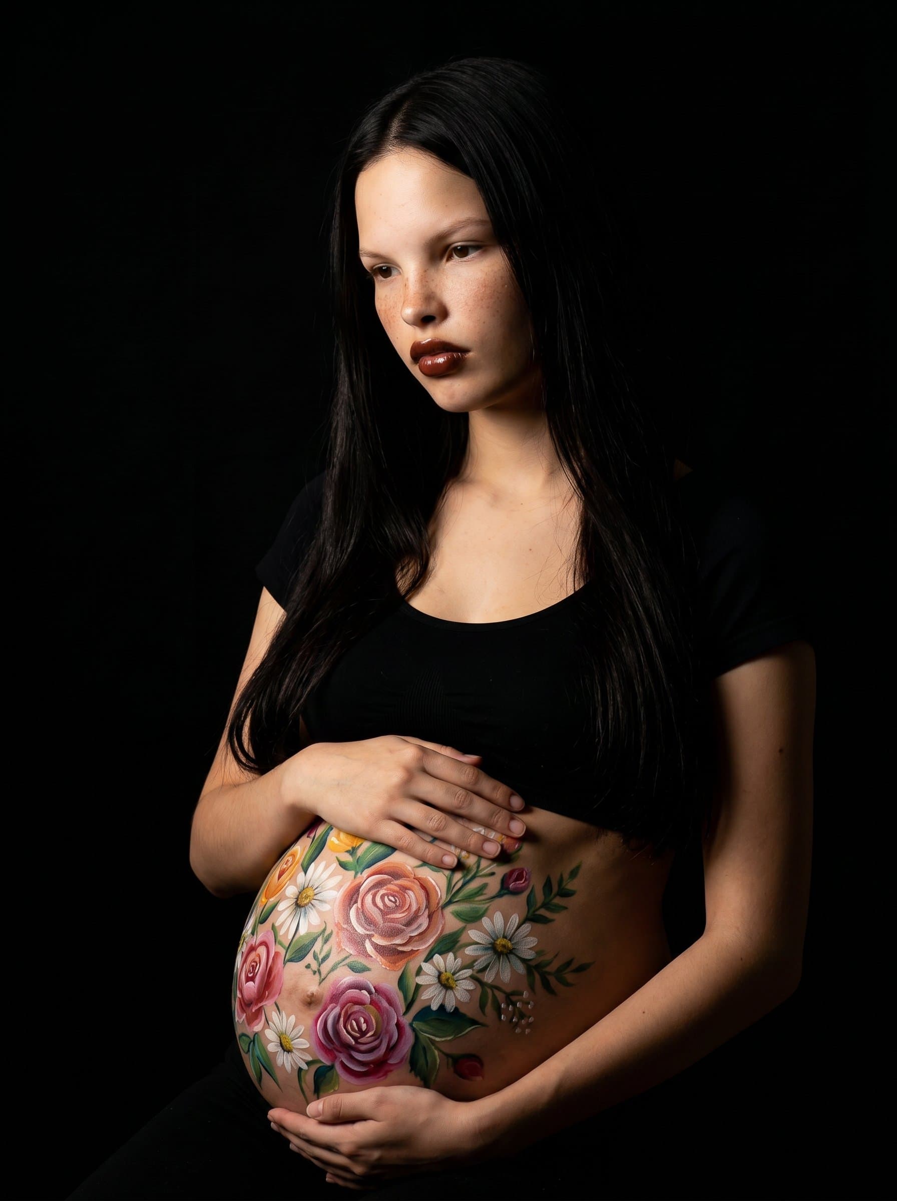 Artistic portrait of pregnant painted with body art flowers on belly, studio black background, dramatic side lighting, creative maternity art photography