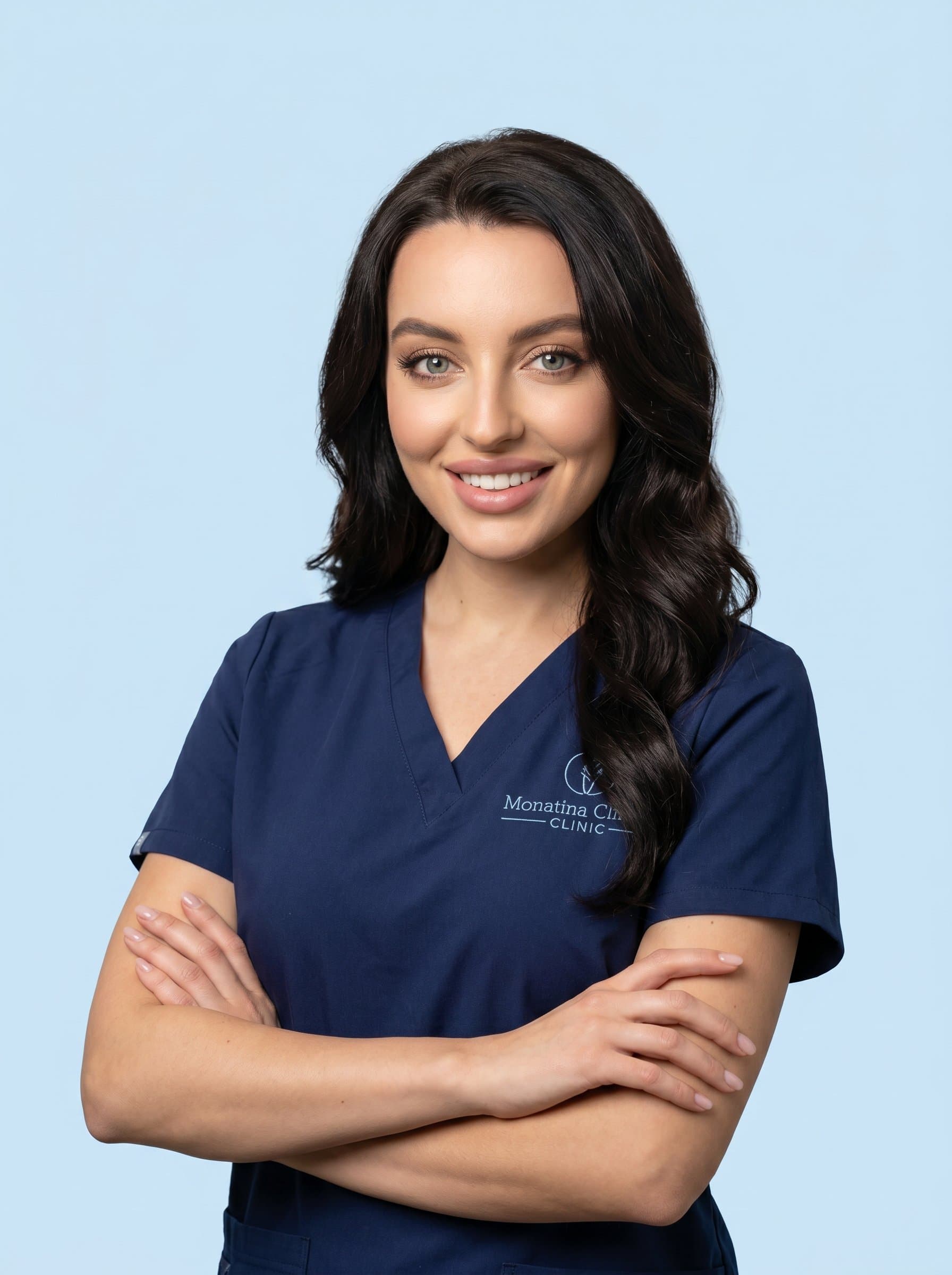 Professional headshot of with arms folded, wearing a neatly pressed scrub top. The backdrop is a soft, pastel color, emphasizing their authoritative yet caring presence, suitable for a clinic's web page.