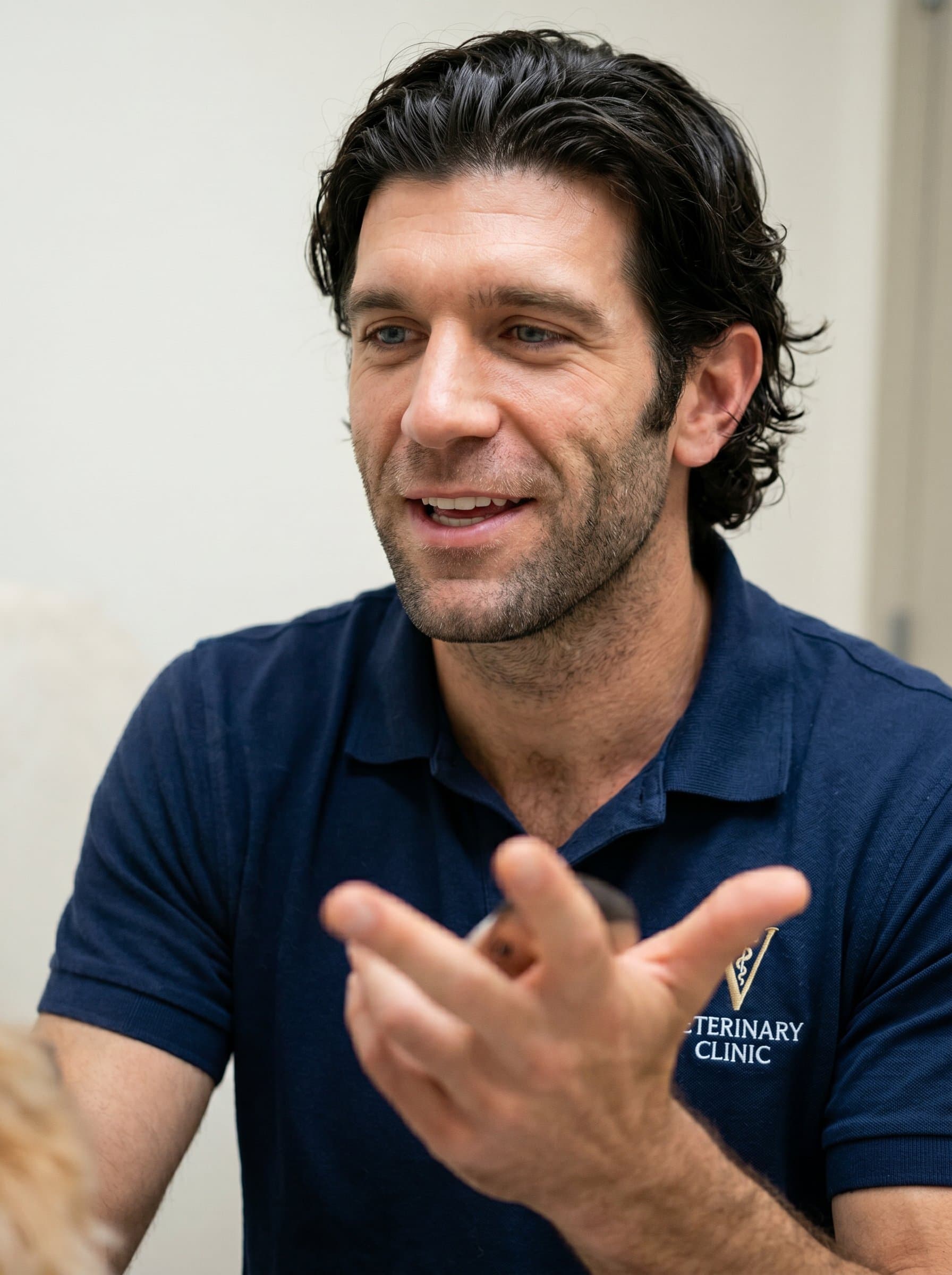 Capture in mid-conversation pose, gesturing towards an unseen pet. Wearing a polo shirt with a clinic logo, they exude friendliness and confidence, with a neutral backdrop enhancing focus on their expression.