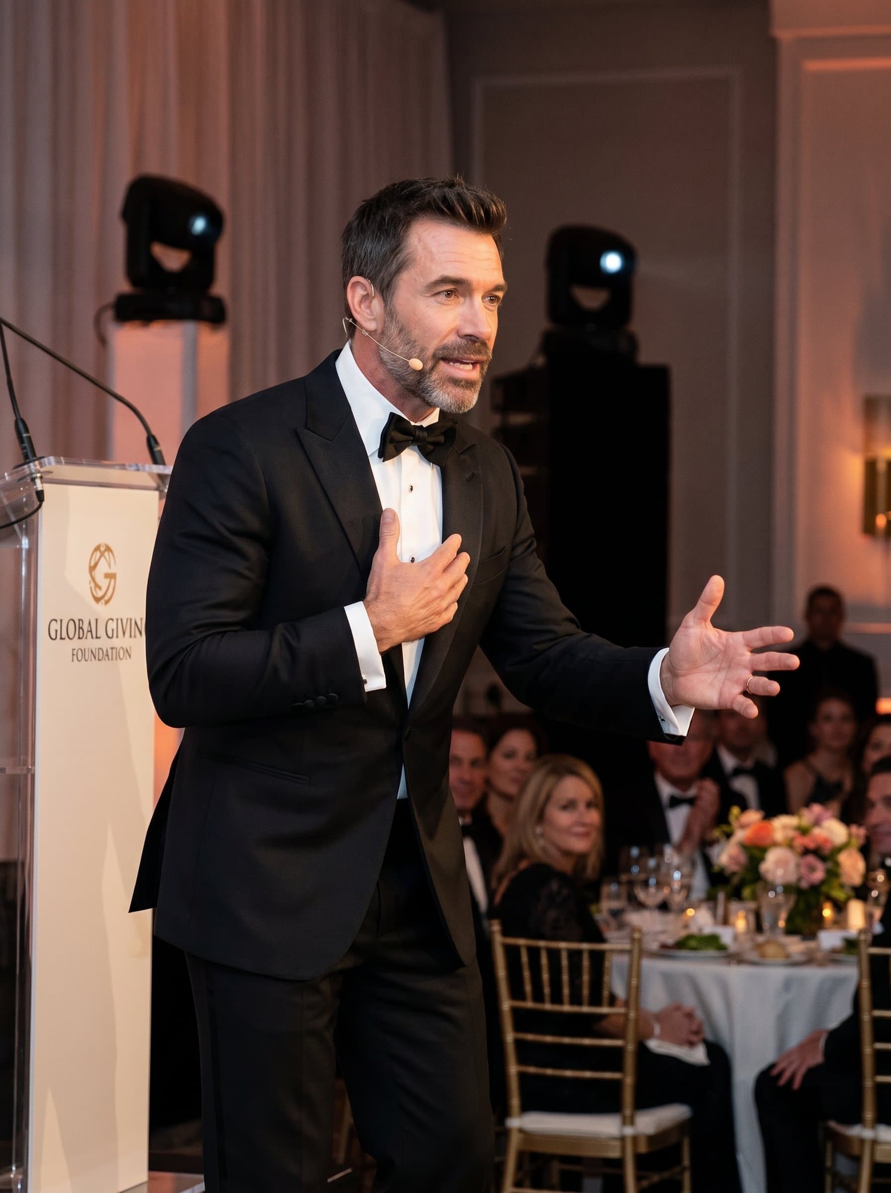 Capture a of a speaker at a charity event, dressed in formal evening wear, using expressive hand movements to convey empathy and action.