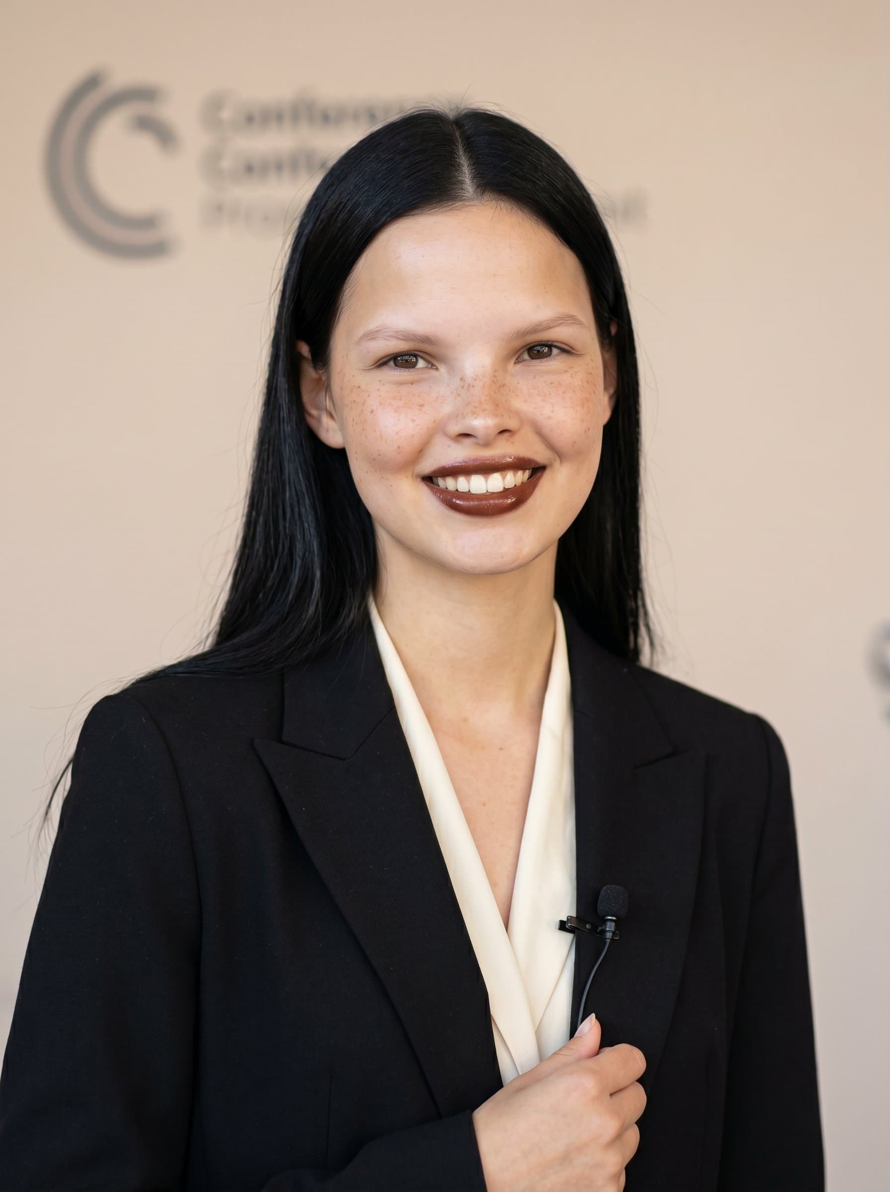 Create a portrait of an articulate speaker with a warm smile, wearing a stylish black blazer. The soft beige backdrop highlights the clarity of the woman's skin and the friendliness of their eyes.