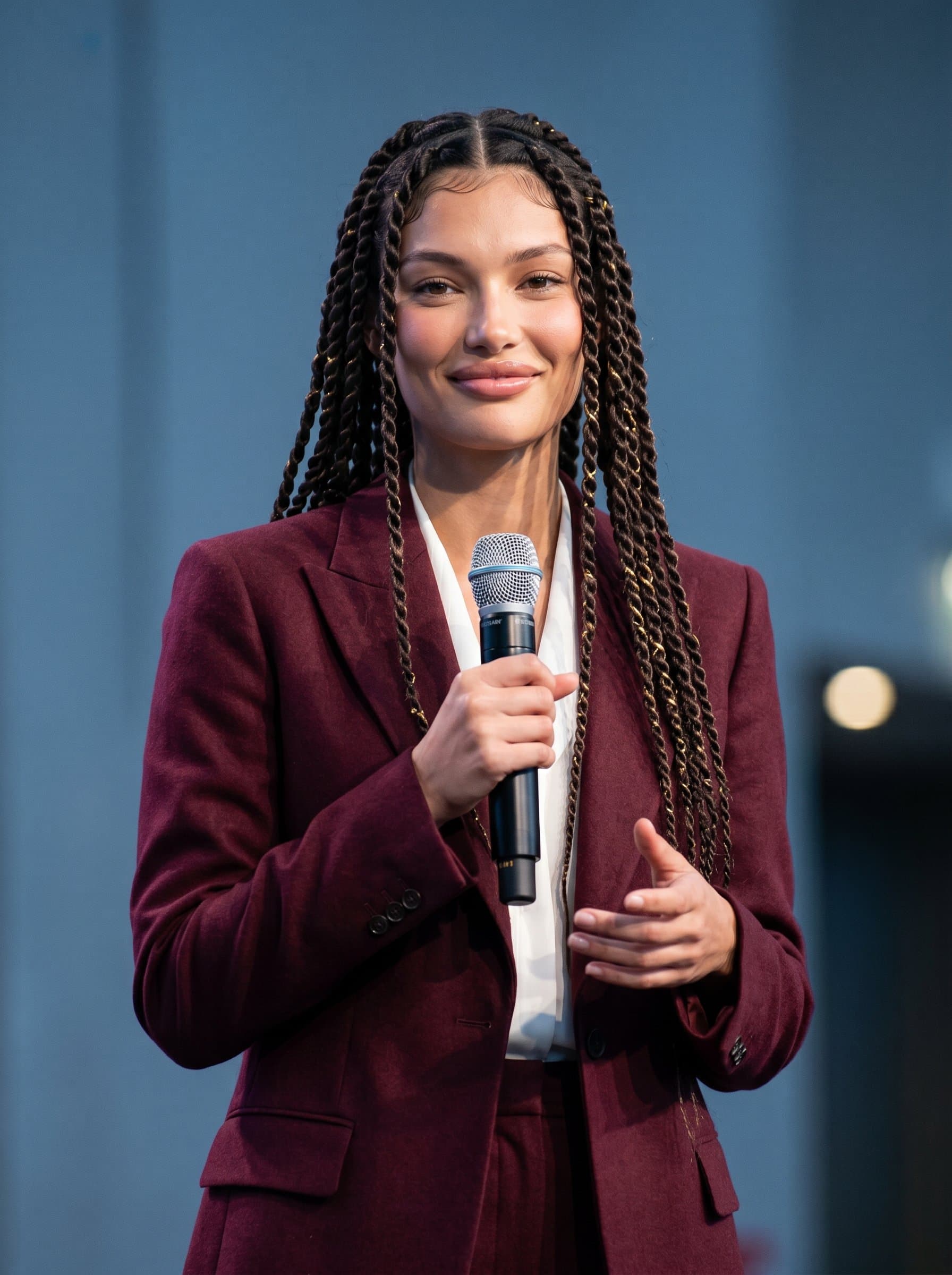 Generate a image of a charismatic speaker with a gentle smile, dressed in a well-fitted burgundy suit. The background is a muted blue, drawing attention to the woman's engaged and friendly gaze.
