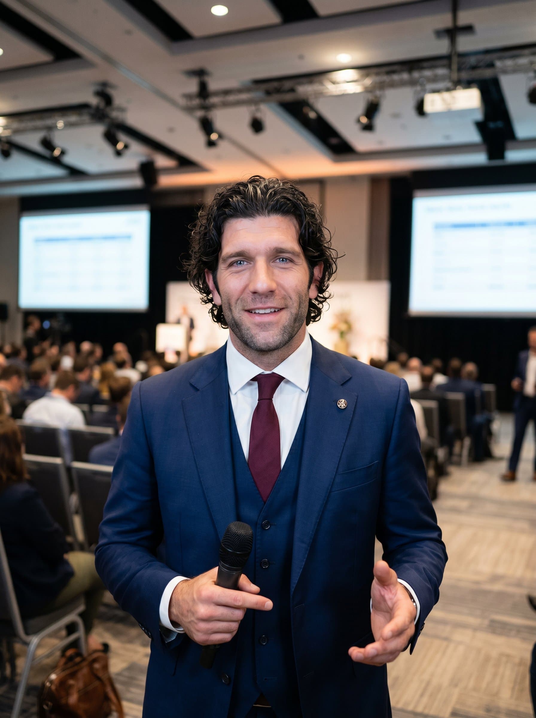 Capture a image of a keynote speaker exuding confidence, dressed in a sleek navy suit. A blurred conference room background subtly suggests the speaker’s environment, focusing on their charismatic expression.