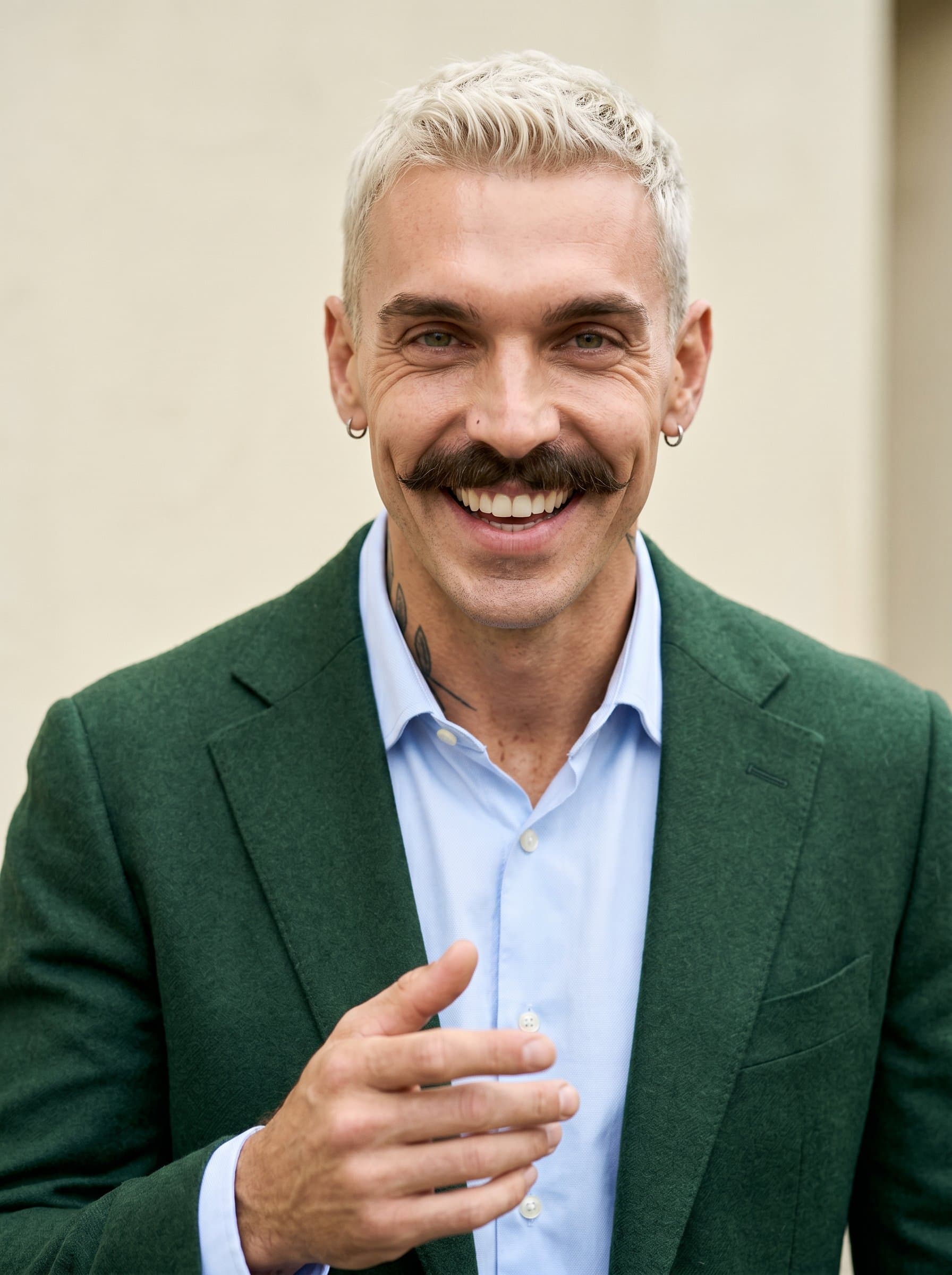 Produce a headshot of a speaker with an expressive face, sporting a tailored green blazer. The background is a muted cream, ensuring full attention on the man’s engaging and inviting look.