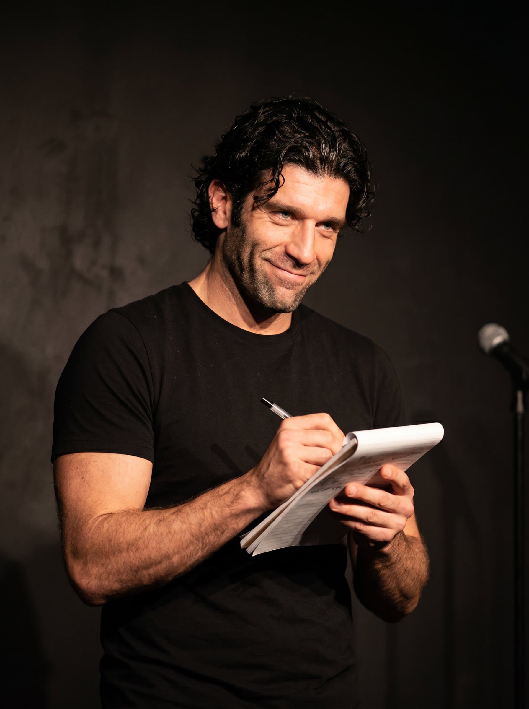 : Comedian holding a notepad, pretending to jot down a joke. Their expression is focused yet playful, set against a minimalistic background to highlight their creative process.