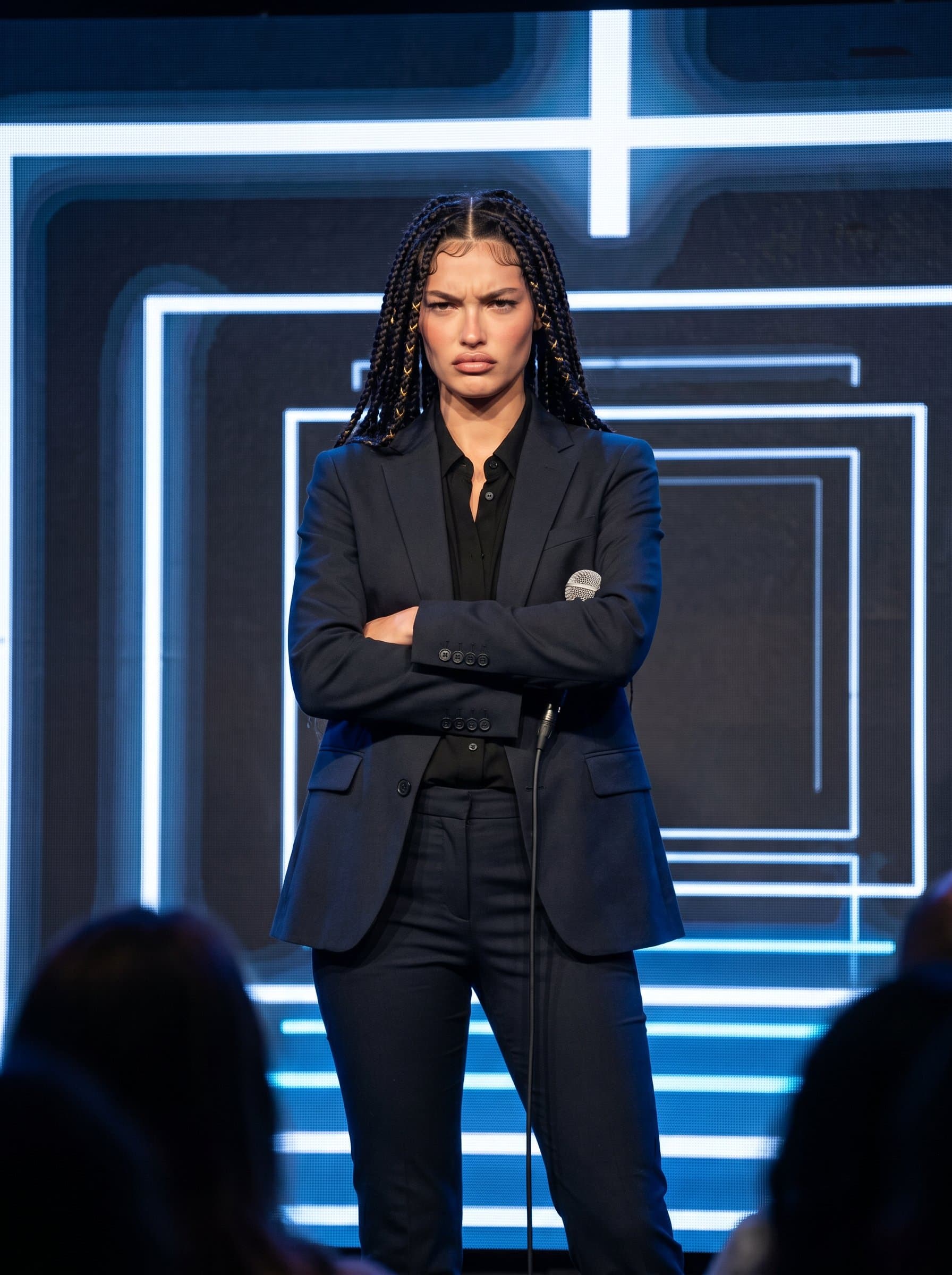 : Comedian delivering a mock-serious expression, arms crossed defiantly. They wear a smart-casual outfit, and the modern digital backdrop adds a crisp, clean aesthetic.