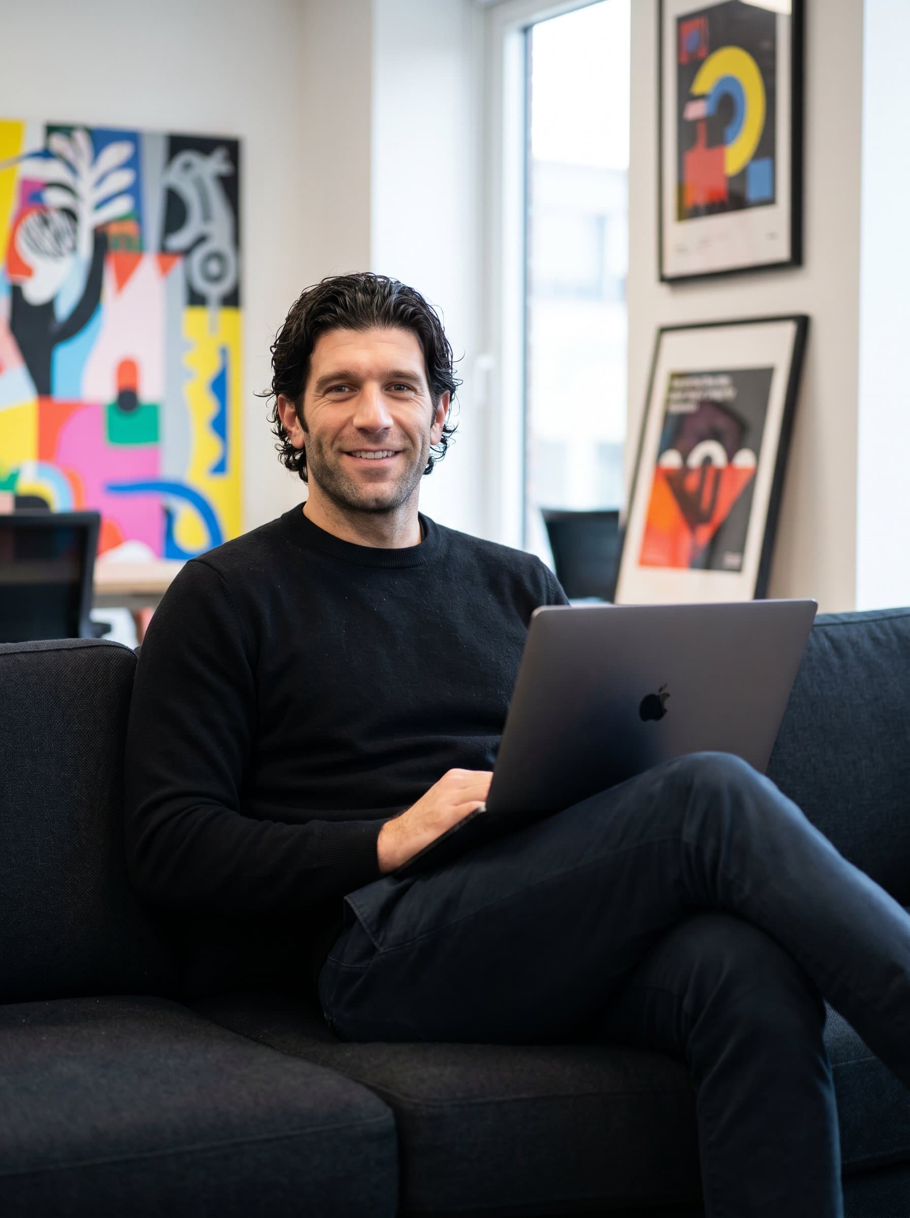 : Portray a confident product manager, casually sitting on a couch with a laptop in their lap. The blurred office artwork behind them conveys a creative and professional work environment.
