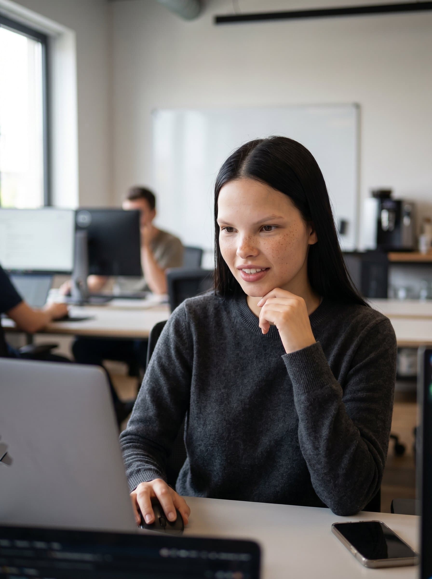 : Capture a product manager in a modern workspace, focused yet approachable, in a neat sweater. Soft lighting highlights their facial features, with blurred office technology subtly visible.