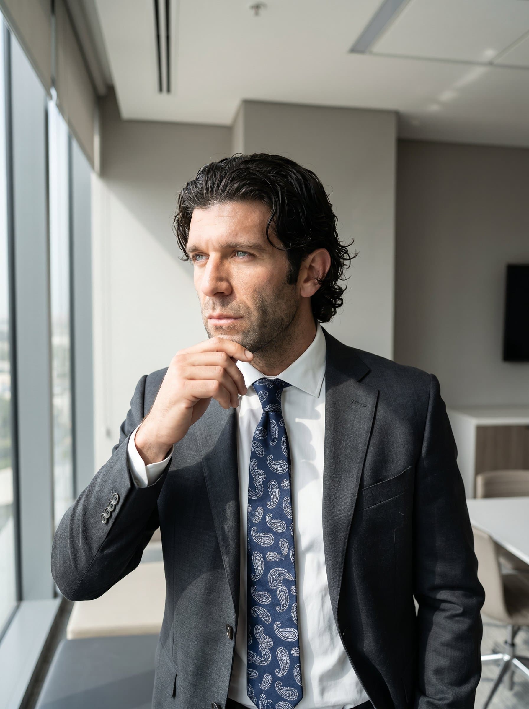 A thoughtful half-turned toward a window with natural light, wearing a stylish necktie. The room is modern with neutral tones, enhancing the impression of reliability and understated elegance.
