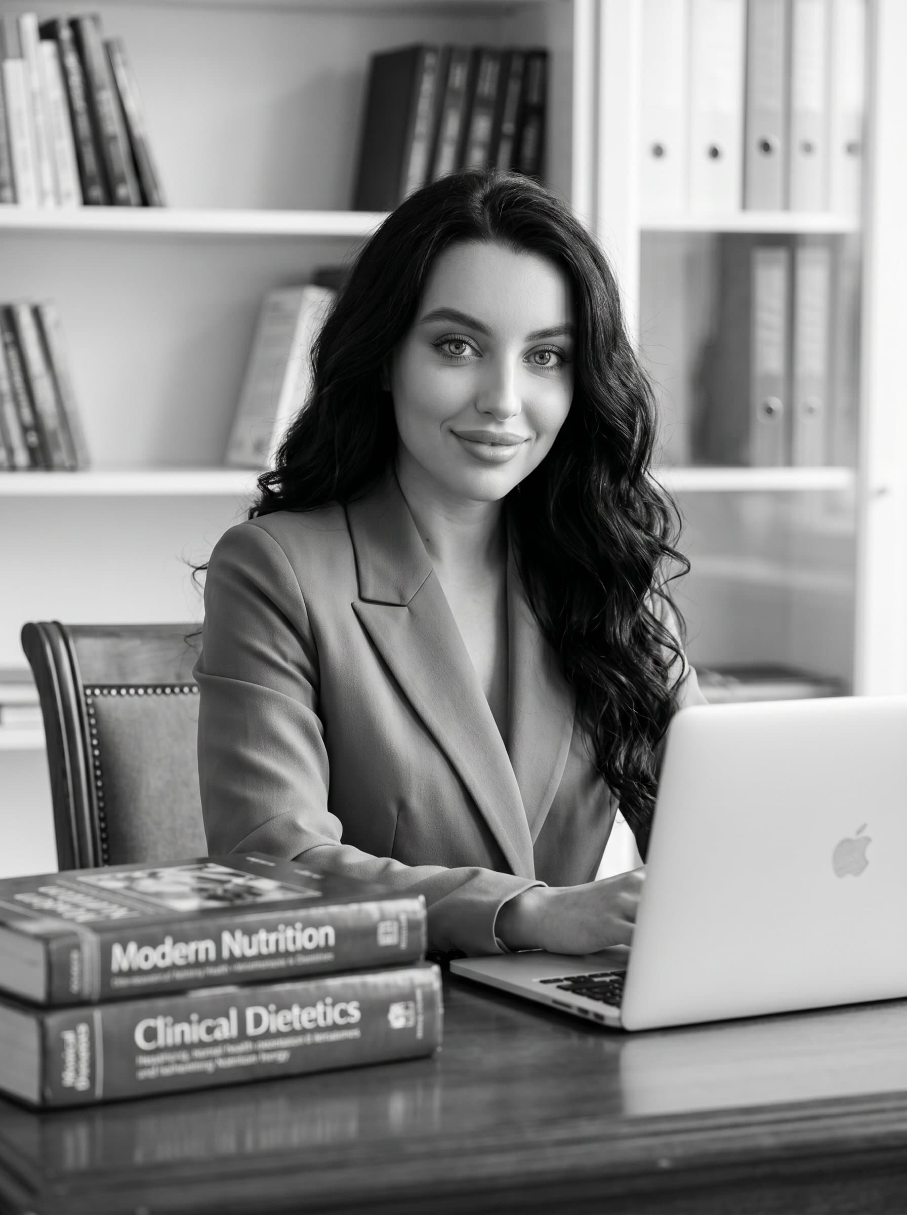 : A monochrome headshot of a dietitian in a classic office. They are seated at a desk with a laptop and nutritional books, exuding a calm and focused demeanor.