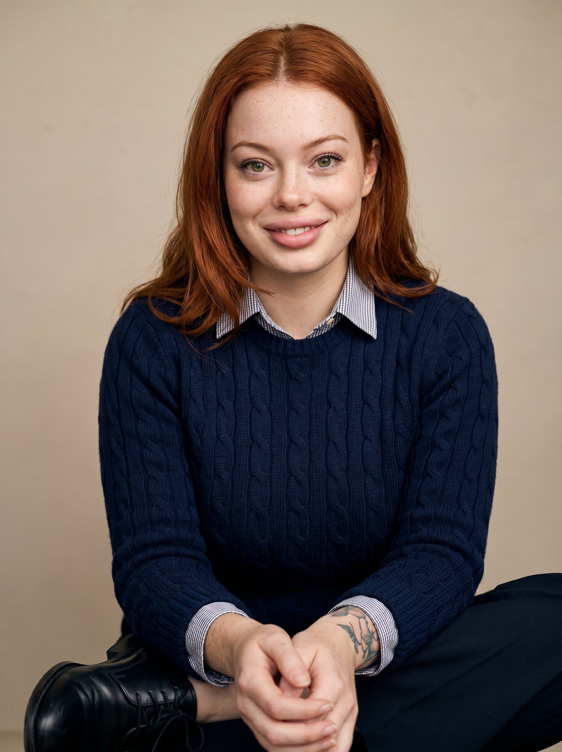 : The chiropractor is captured in a neutral studio setting, wearing a classic sweater and dress shoes, with a composed posture and a direct, understanding look that conveys trust and expertise.