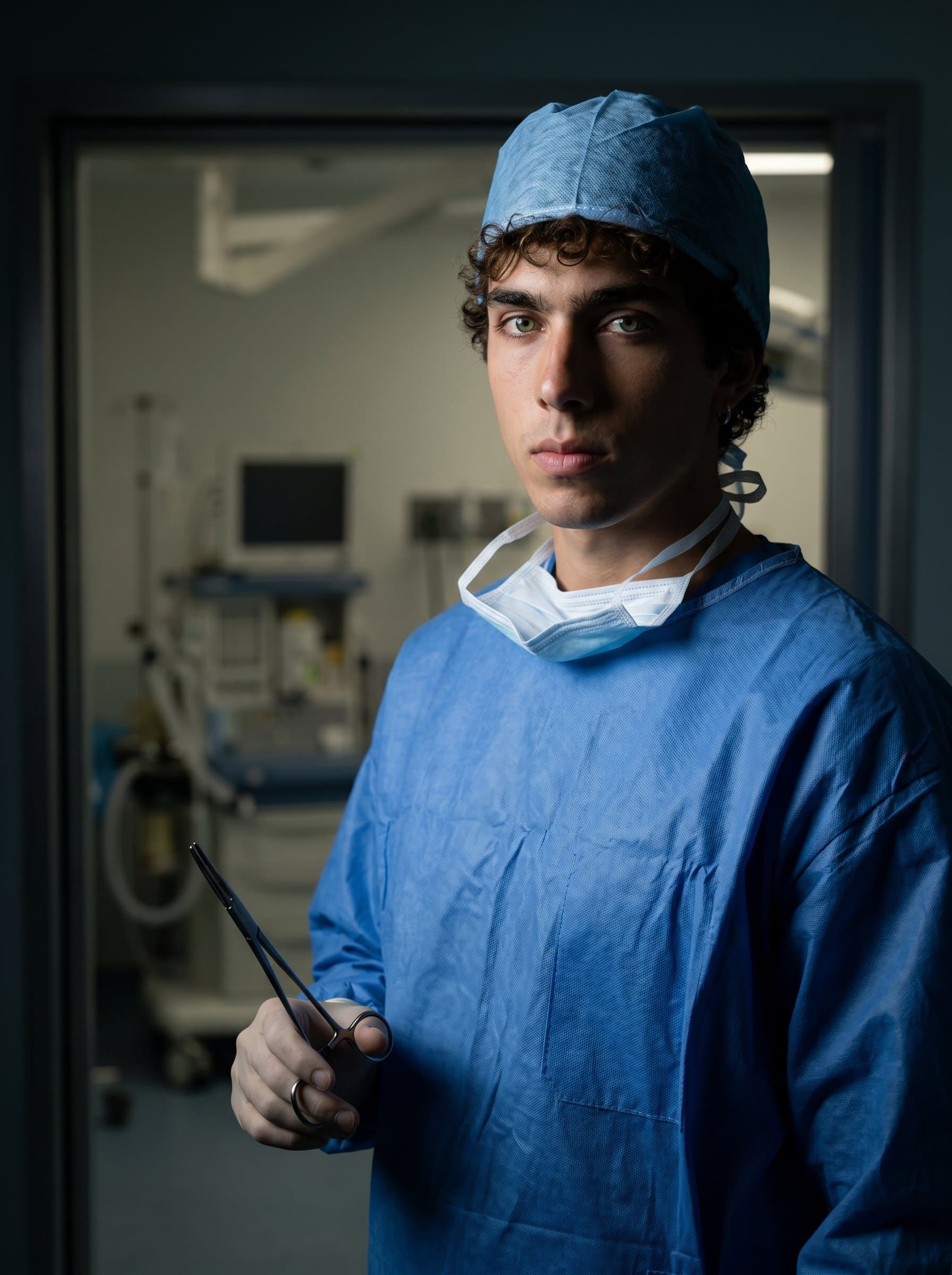 surgeon, holding a surgical instrument, standing in a dimly lit prep room. The shadows and concentrated expression highlight seriousness and readiness, ideal for a professional profile focused on precision and commitment.