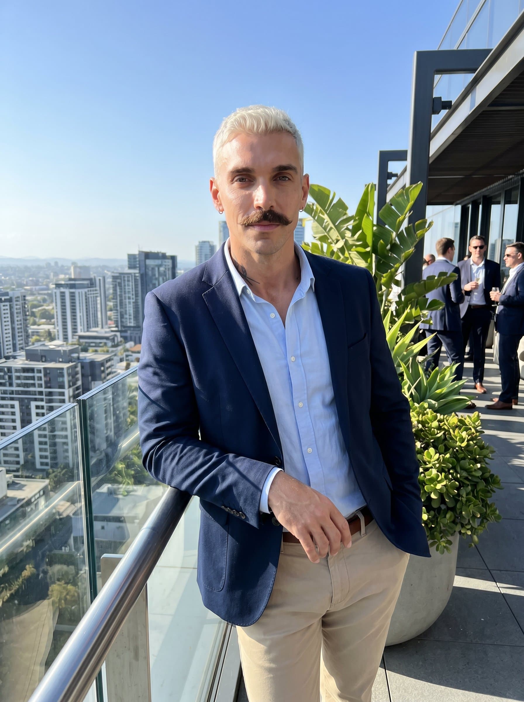 On a sunlit terrace with a skyline view, leans slightly on the railing, exuding calm confidence. Their smart, casual look and serene expression make them appear both professional and approachable.