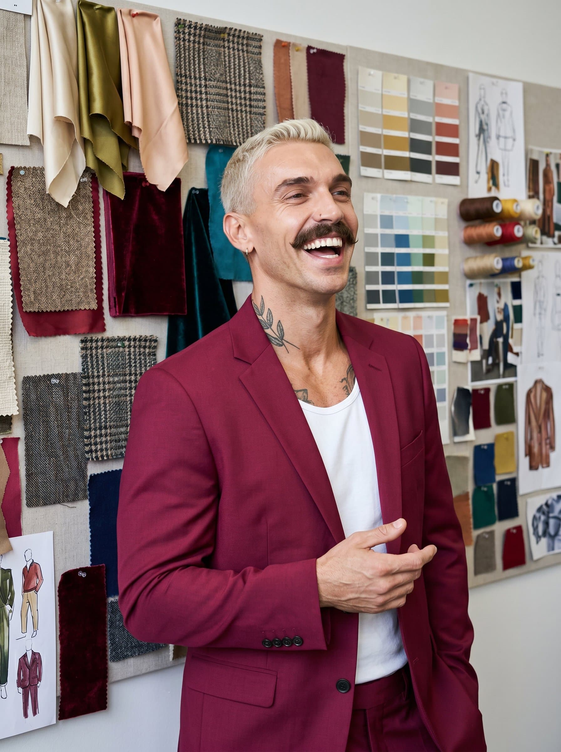 captured mid-laugh, wearing a vibrant maroon blazer, standing before a wall filled with color swatches and fabric samples, embodying a dynamic and friendly disposition.
