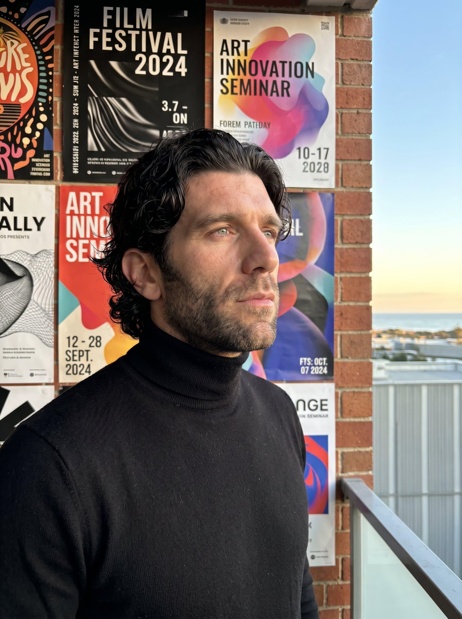 gazing thoughtfully towards the horizon, dressed in a stylish black turtleneck, with a wall adorned with artistic event posters behind, reflecting an eye for detail and an innovative spirit.