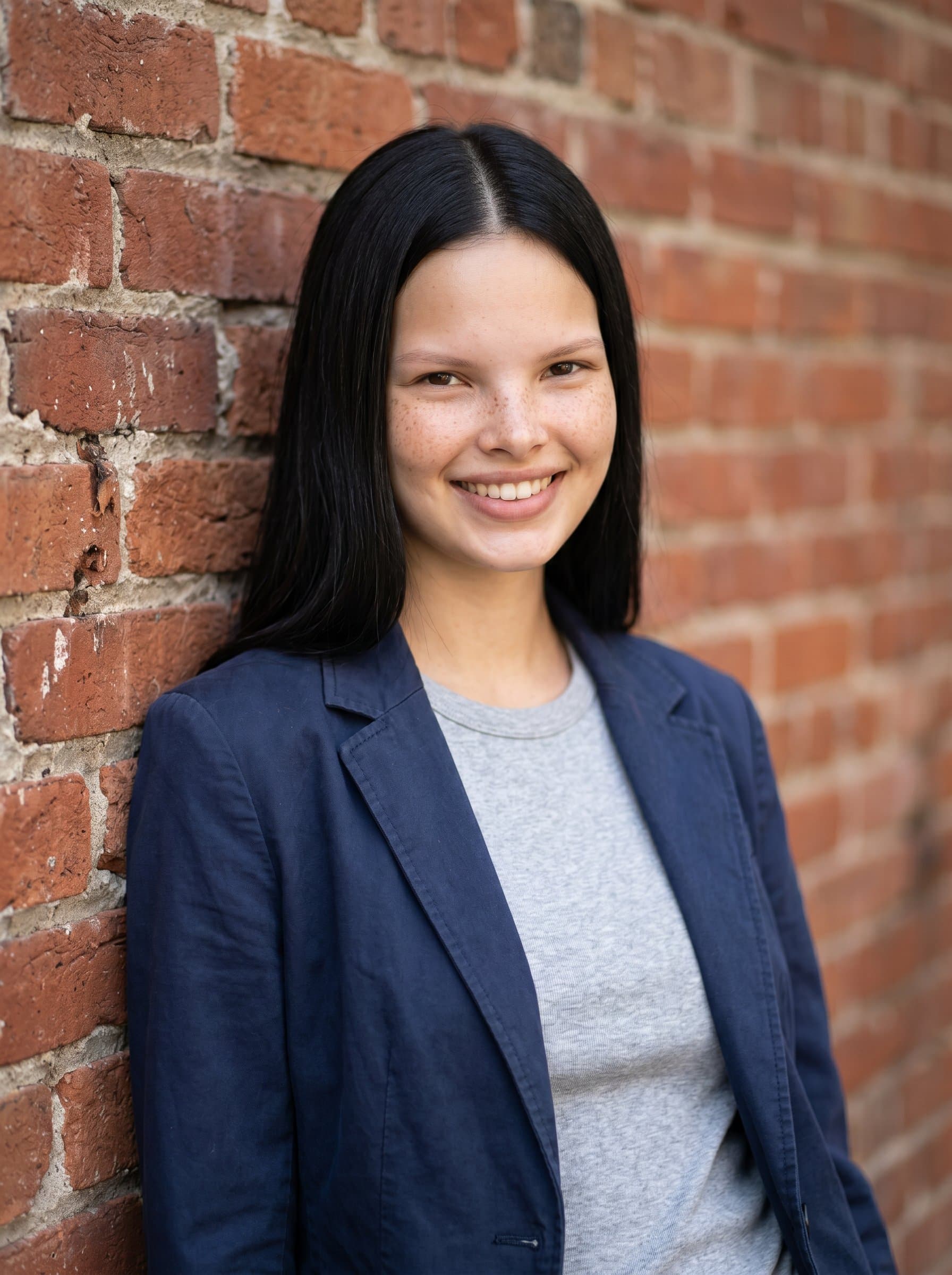 Formulate a headshot for a casual-themed mortgage broker, leaning against a rustic brick wall, wearing a relaxed blazer and t-shirt, highlighting approachability and down-to-earth professionalism.