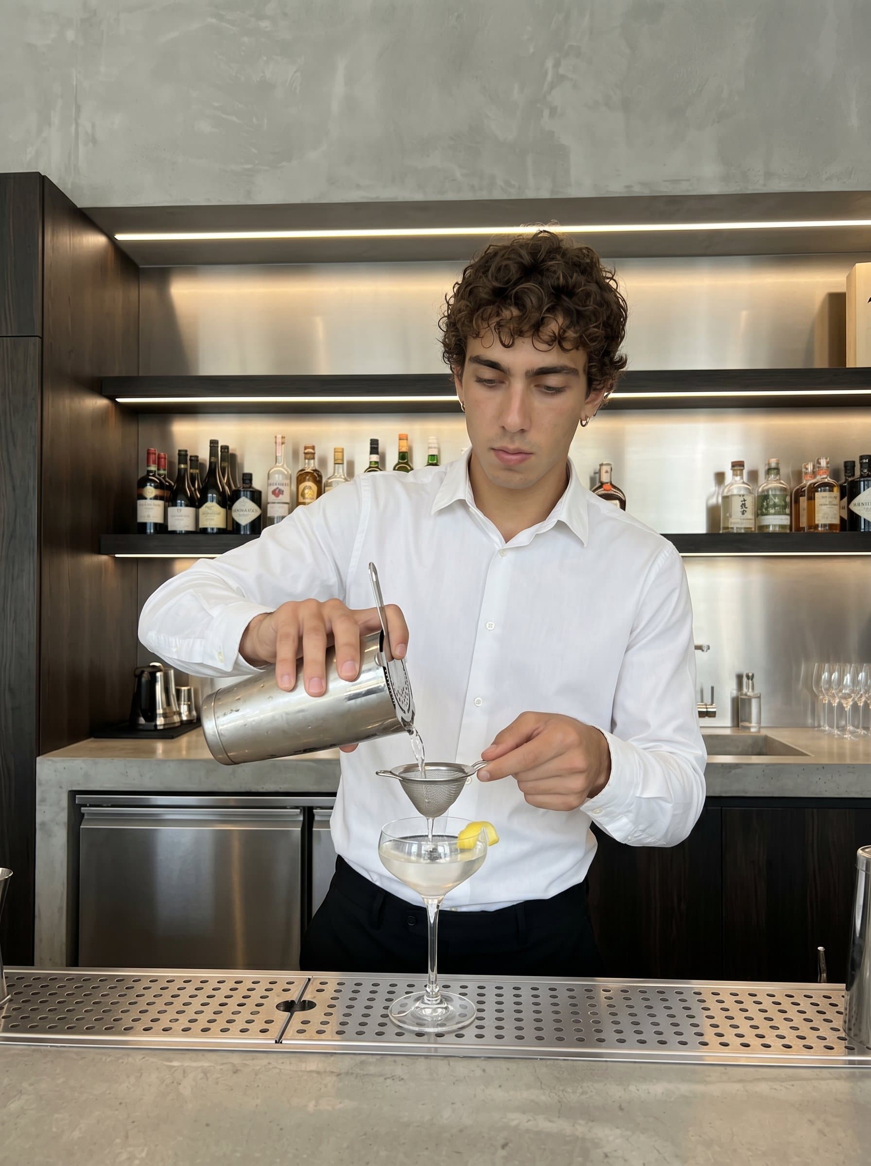 : Capture a bartender with a minimalist style, dressed in a plain white shirt and dark pants, skillfully preparing a cocktail against a sleek, modern bar backdrop with clean lines and subtle lighting.