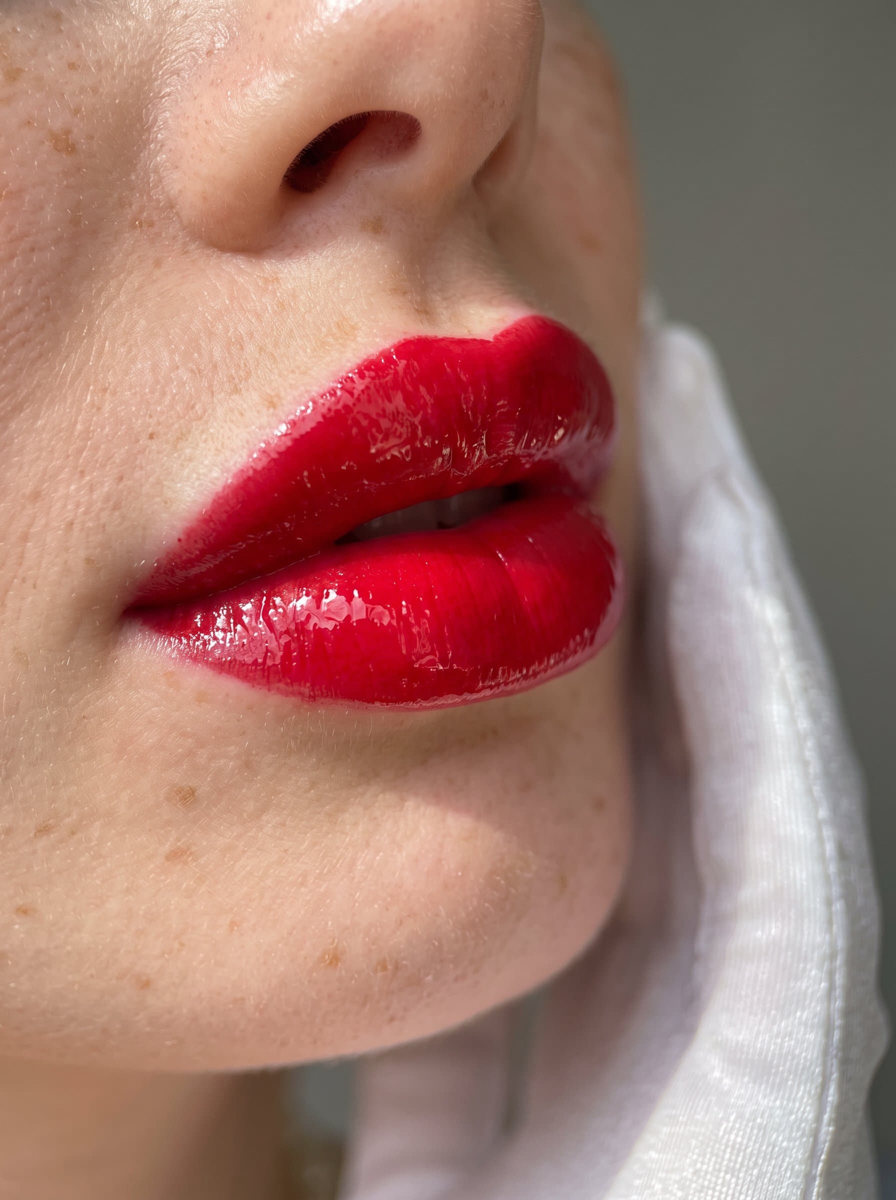 Capture a focusing on bold, glossy lips. The photograph is set against a muted grey background, allowing the vivid lip color to stand out in the natural sunlight.