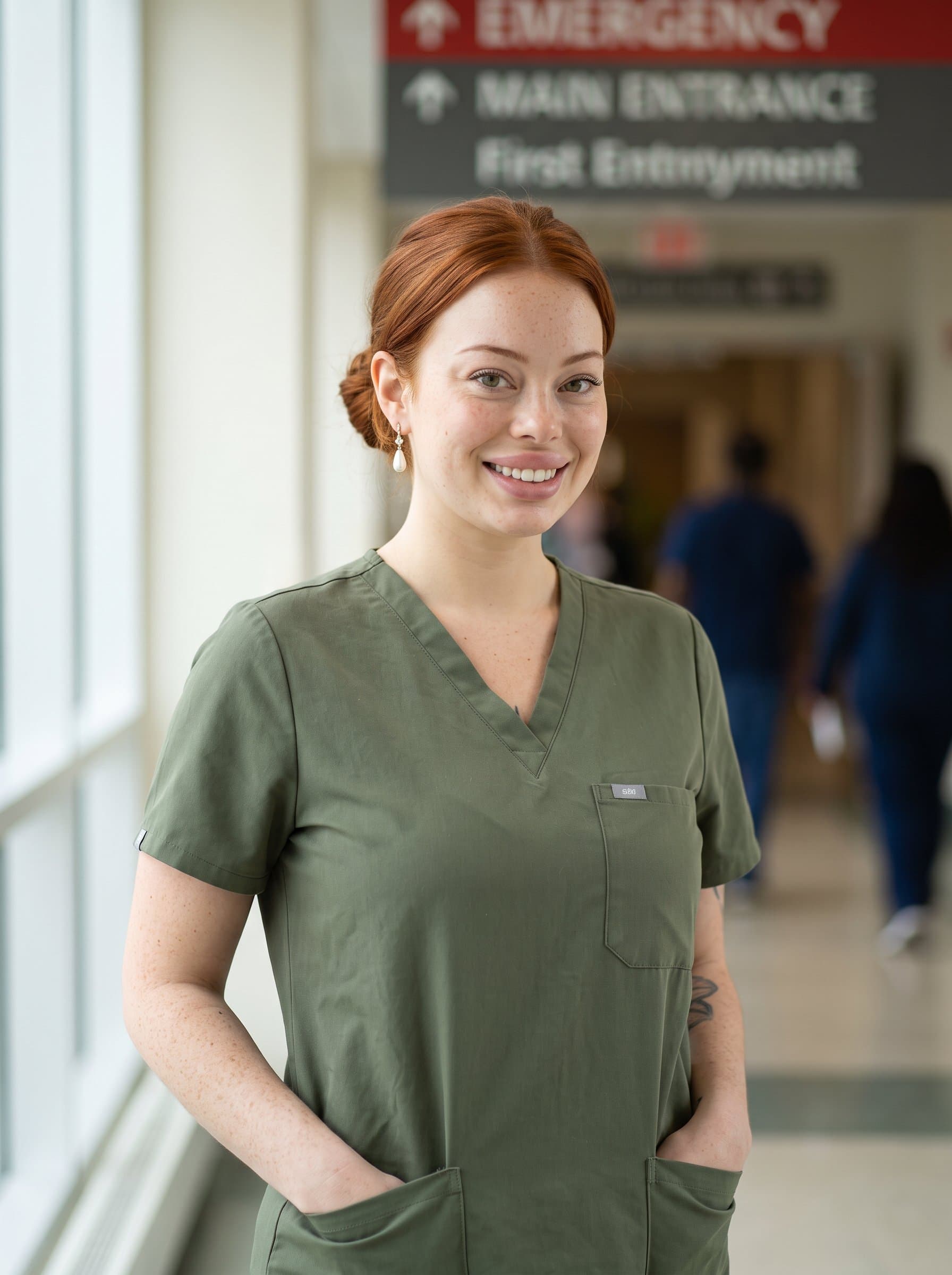: Portrait of a medical professional in olive green scrubs, with hands in pockets, standing in front of a softly defocused hospital sign. Their relaxed posture conveys trust.