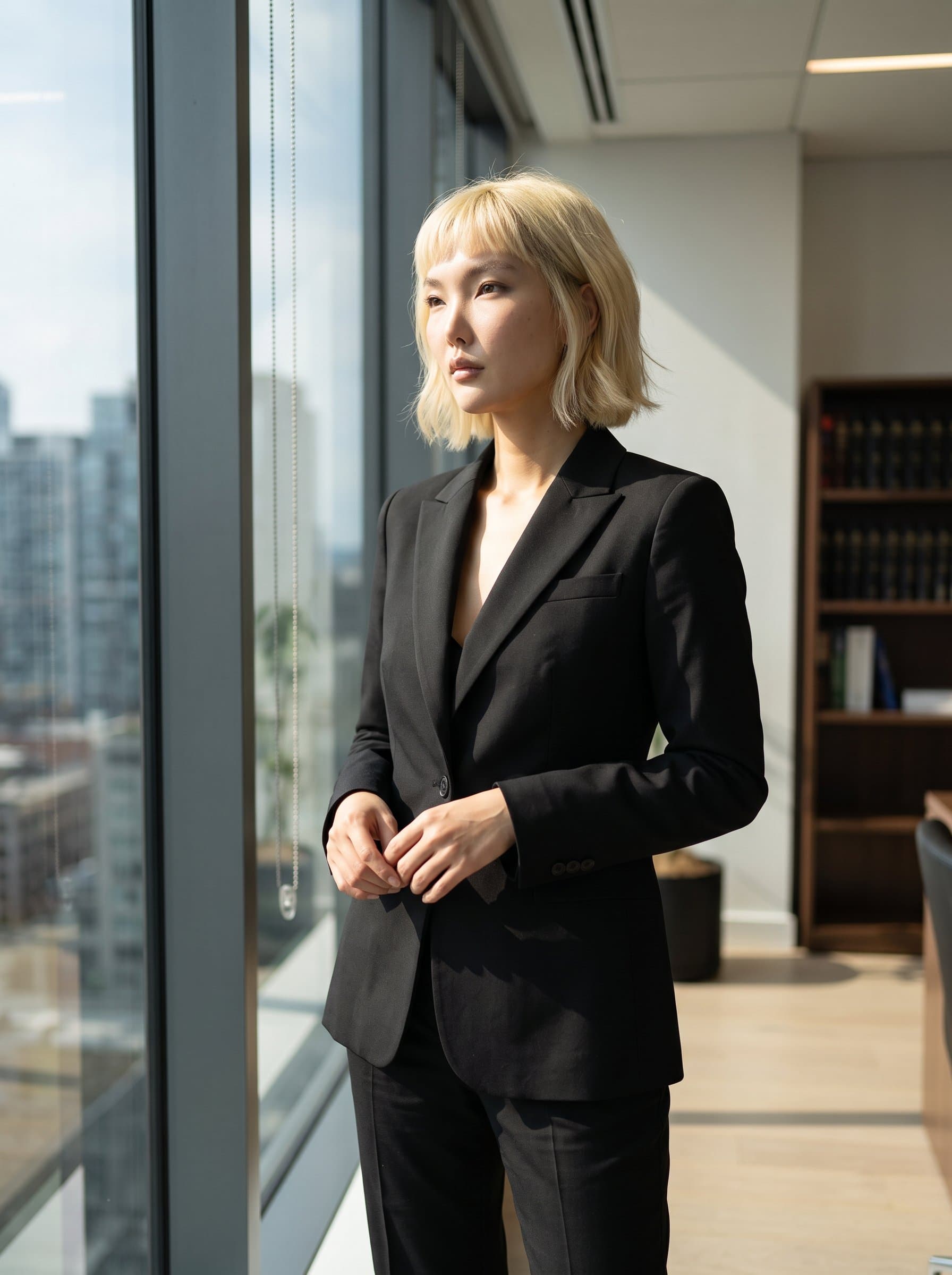 A of a legal professional standing next to a tall window, with natural sunlight partially illuminating their structured suit, capturing a moment of reflection and confidence.