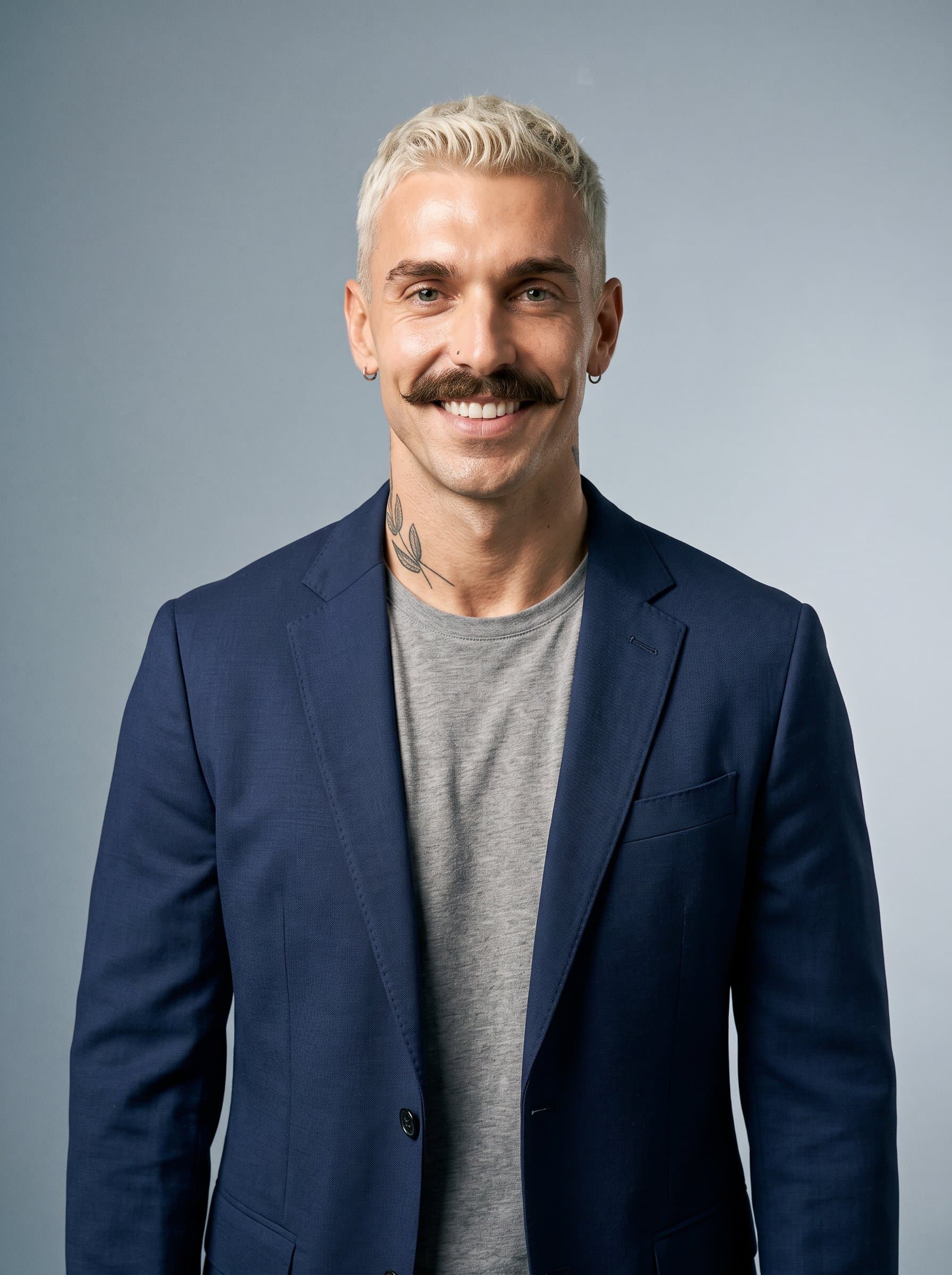 A media consultant with an expressive face and a neat hairstyle, dressed in a casual blazer, looks directly at the camera. enhances their confident presence against a neutral blue-gray backdrop.