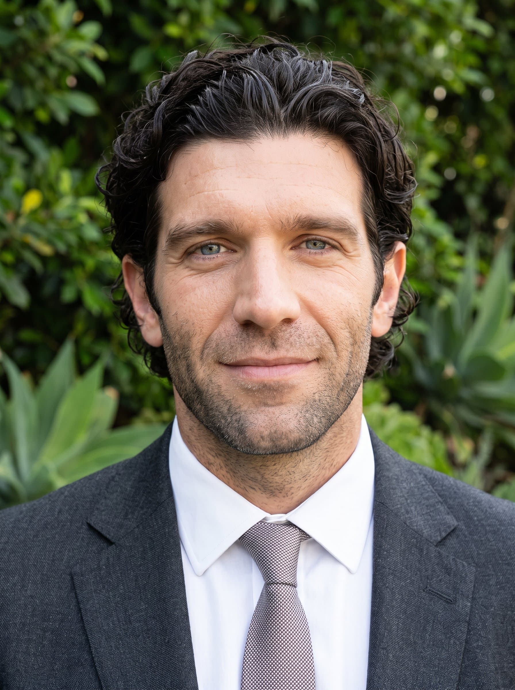 : Generate a headshot of a real estate professional set against a backdrop of green foliage, wearing business attire. The gentle smile and direct gaze suggest trustworthiness and readiness to assist clients.