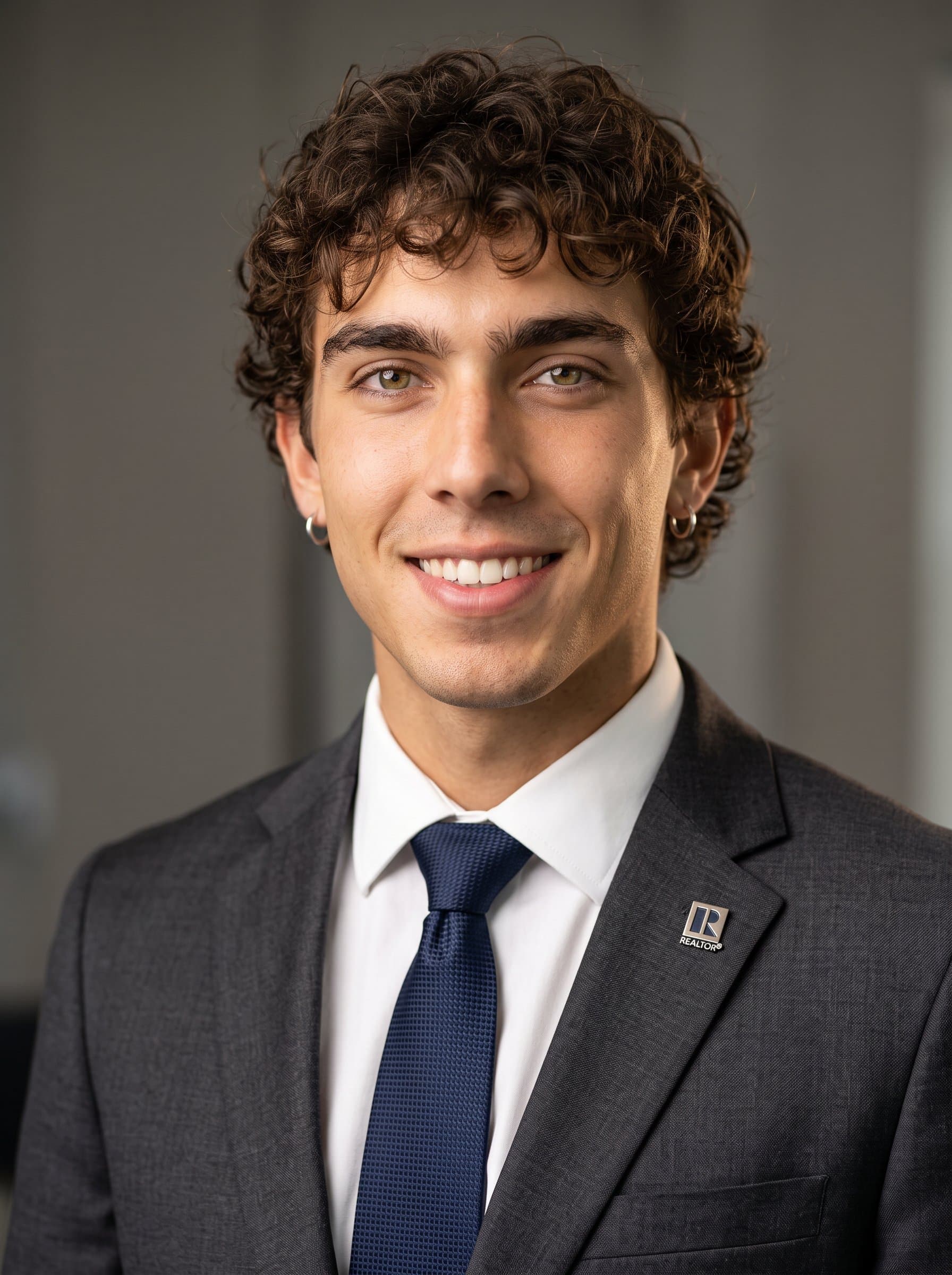 : Frame a headshot of a real estate professional against a neutral background with soft lighting. The man, dressed in business attire, has a calm smile and an inviting expression.