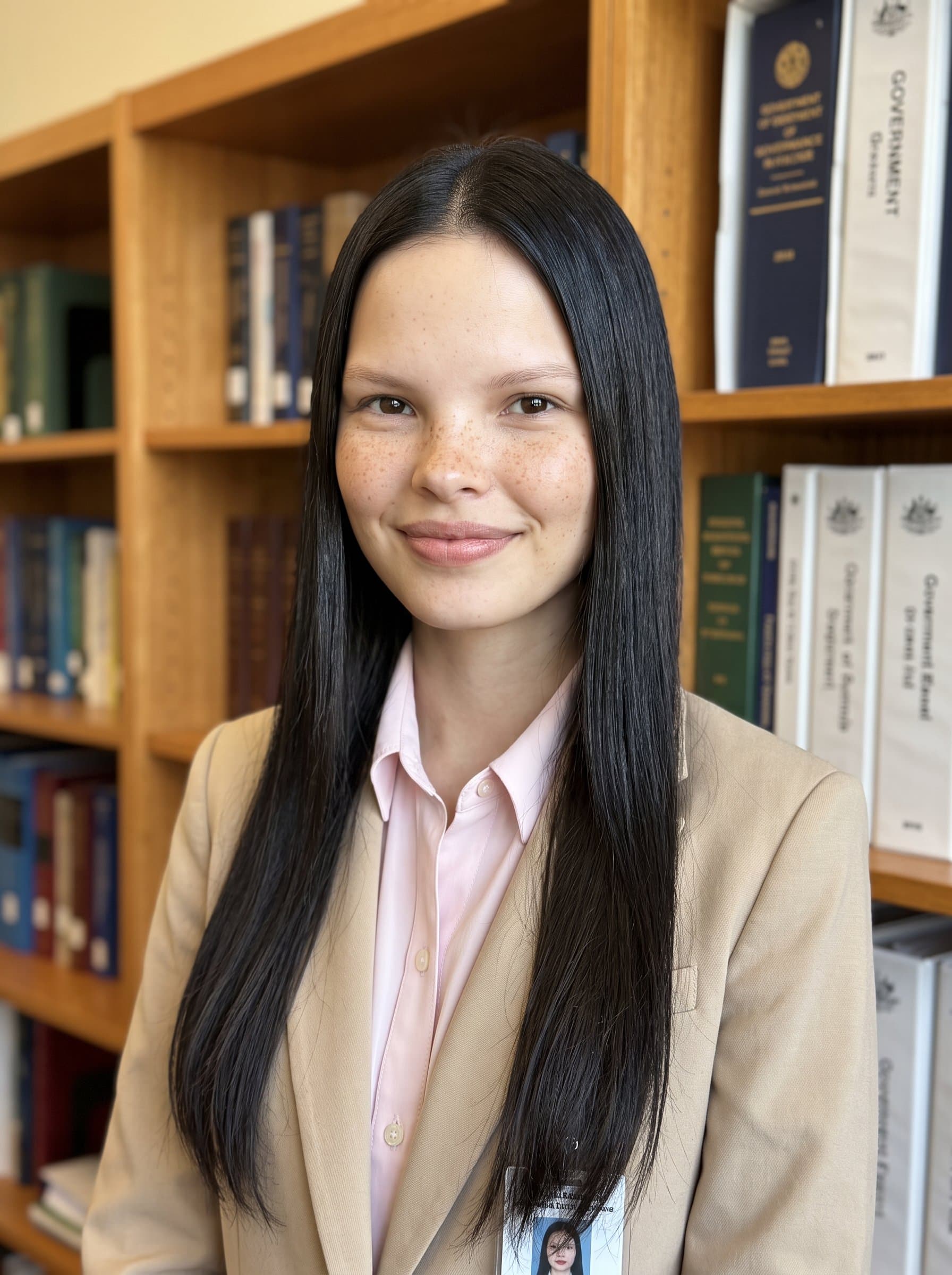 : Picture a government employee against a backdrop of bookshelves, wearing a beige blazer with a pastel-colored shirt, smiling softly, ideal for a professional and approachable profile image. Photorealistic.