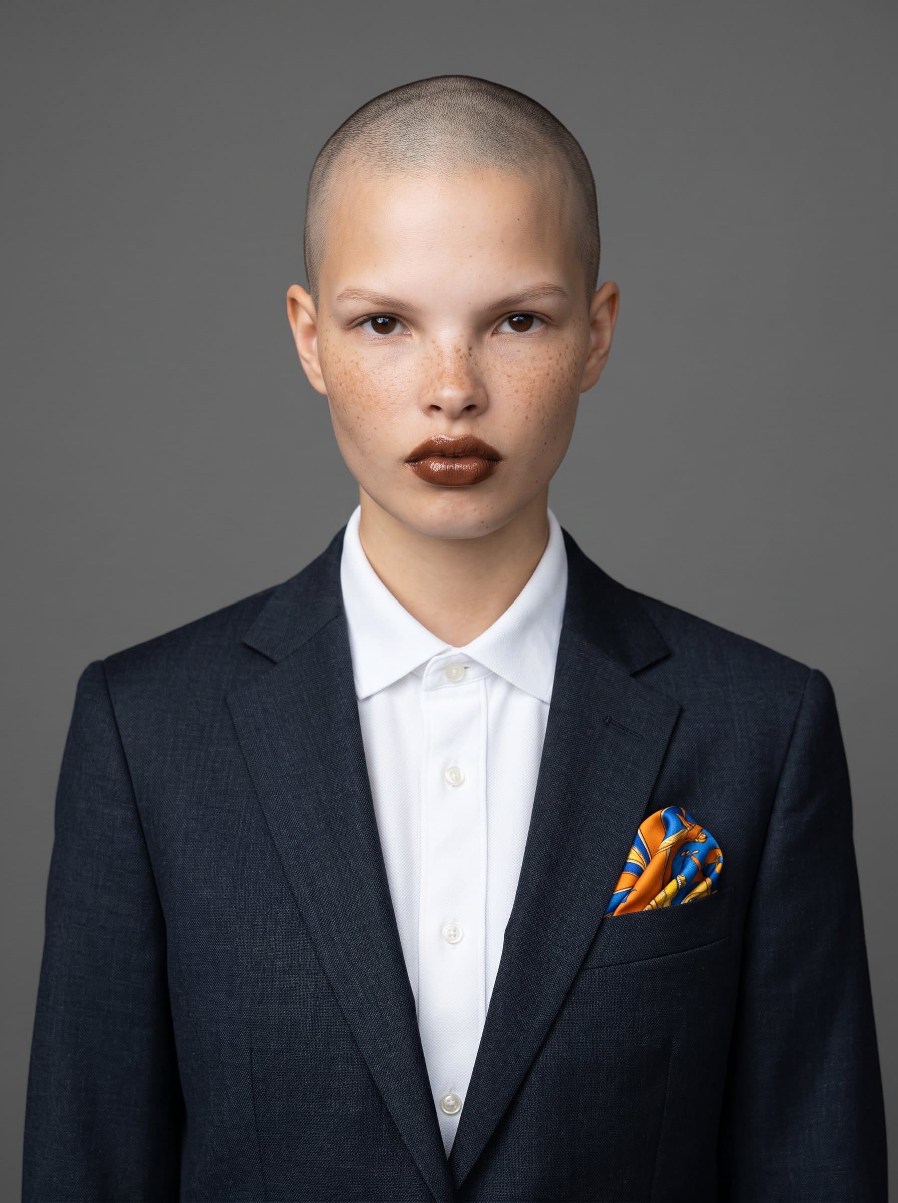 : Photo of a modern consultant with a shaved head, dressed in a dark suit with a vibrant pocket square, standing before a simple gray backdrop, highlighting a calm and assured presence.