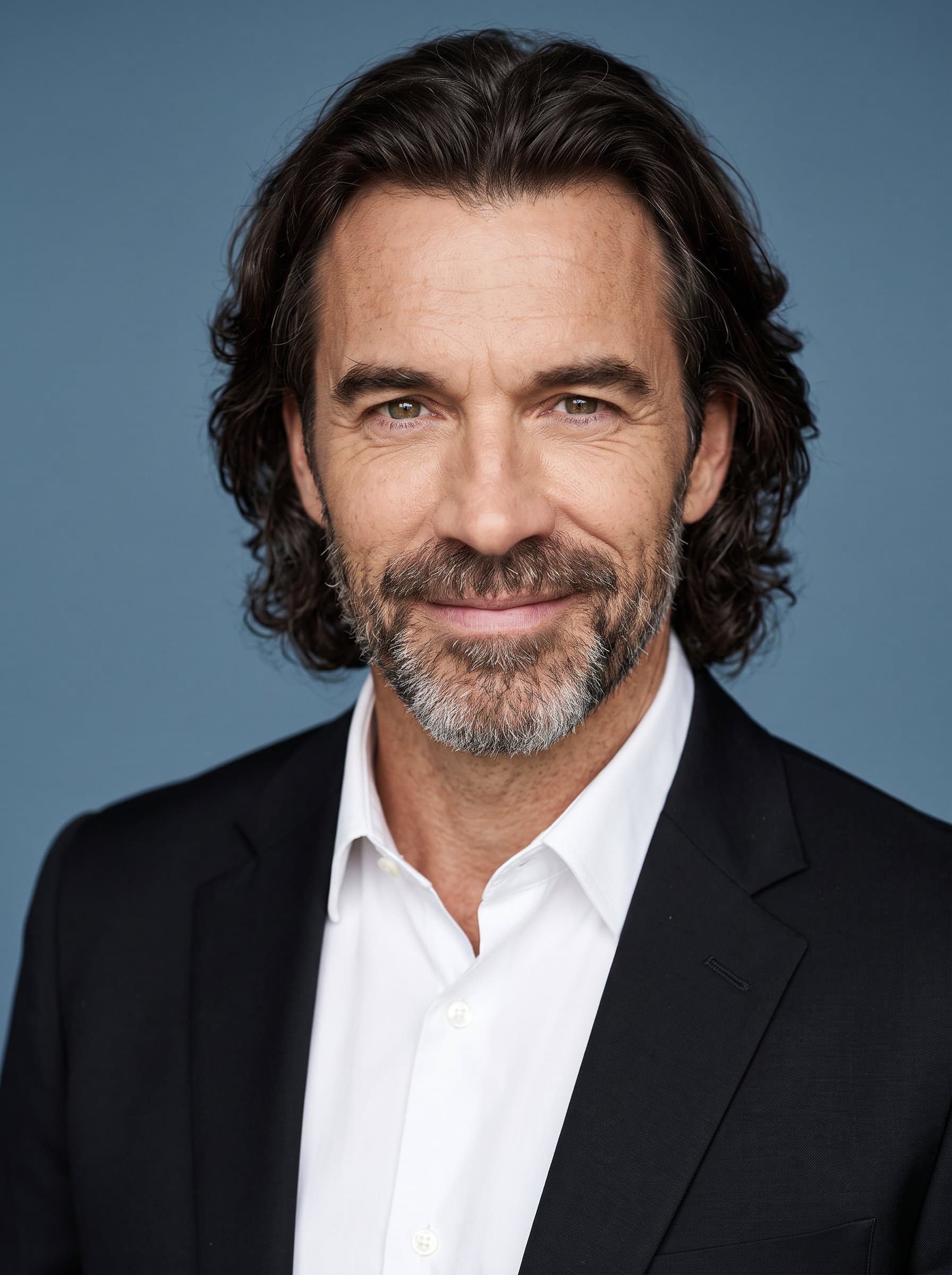 : Headshot of a consultant with long hair, dressed in a classic black blazer and white shirt, captured against a muted blue backdrop, showcasing keen insight and a welcoming expression.
