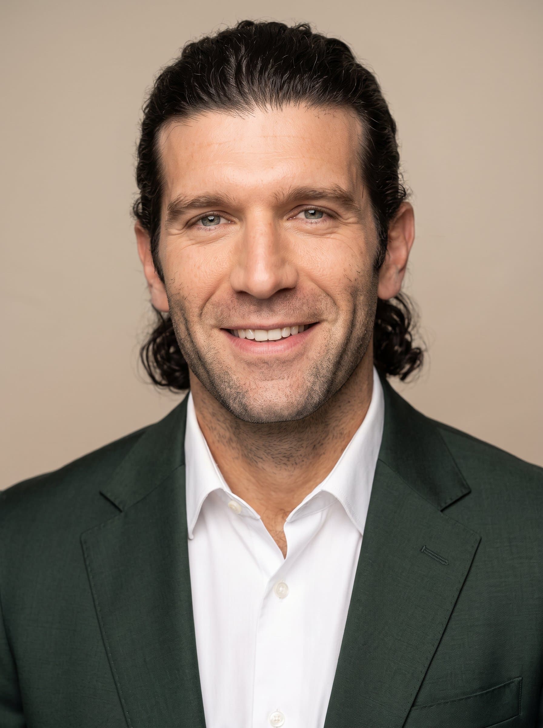 : Headshot of a consultant with a ponytail hairstyle, in a dark green suit and crisp white shirt, captured against an understated beige background, exuding confidence and a friendly disposition.