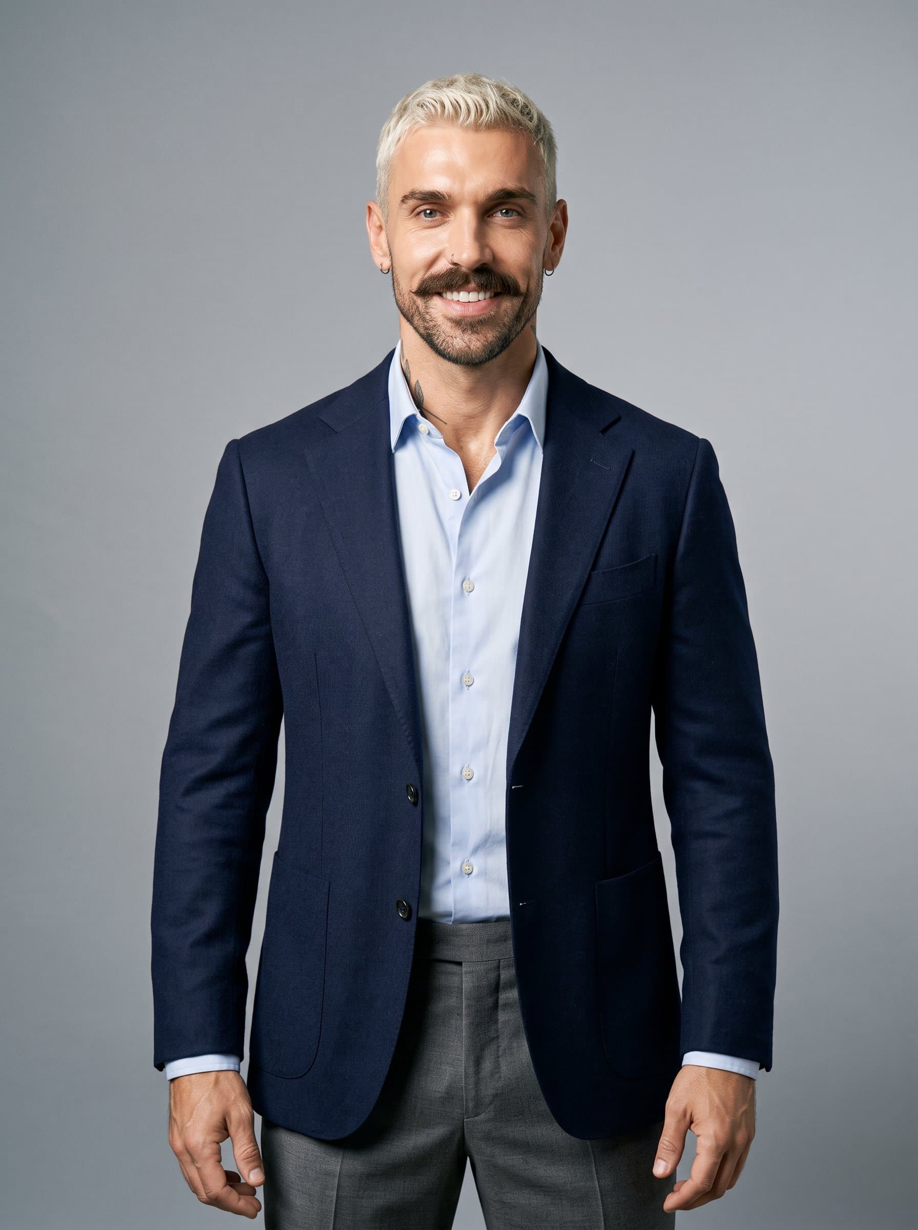 young professional with a closely trimmed beard and short hair, wearing a modern blazer. Background is a sleek, neutral grey, highlighting approachability and a dynamic, professional energy.