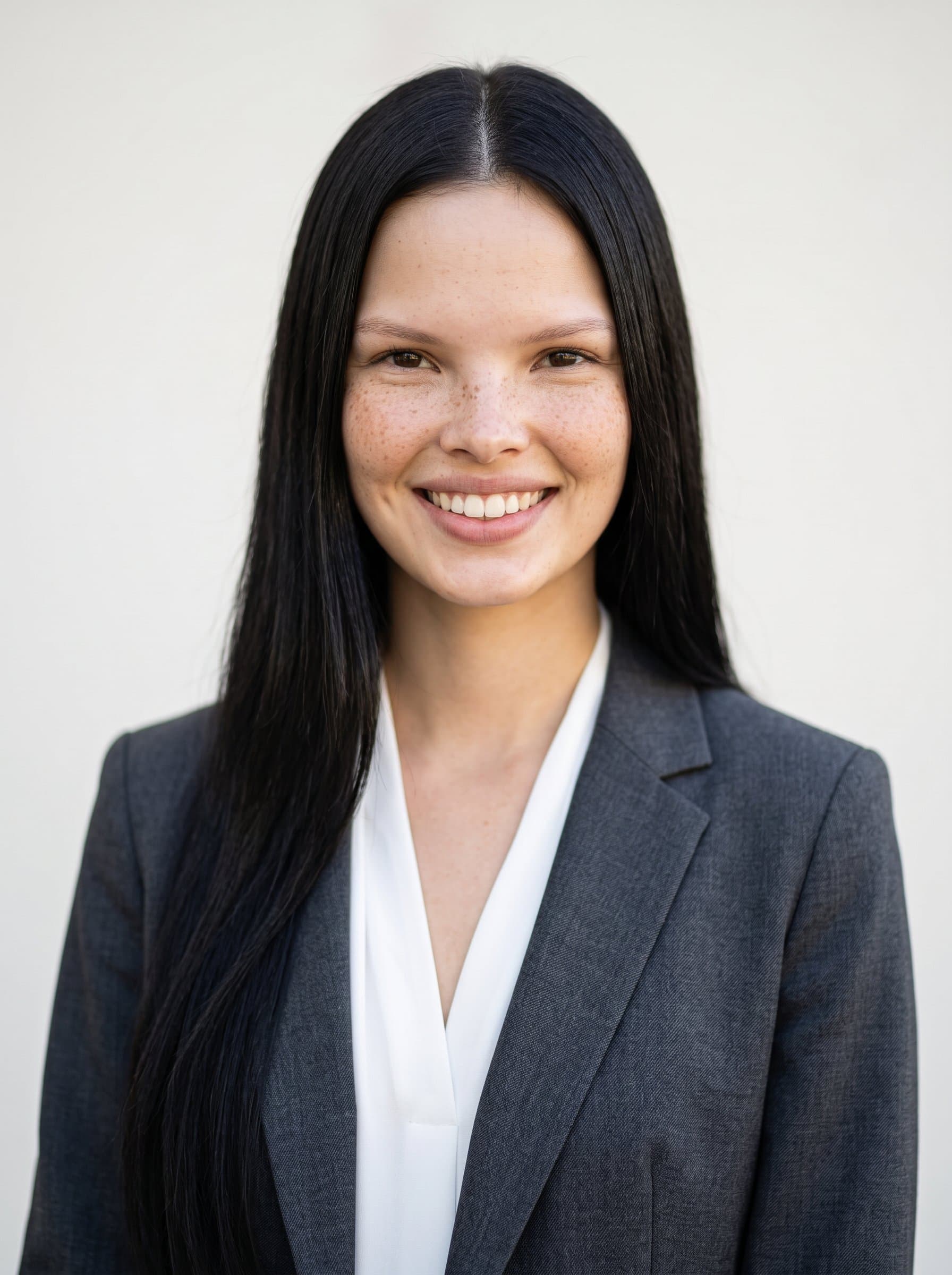 : Design a university faculty headshot featuring a woman in a charcoal suit, set against a subtle eggshell background. Capture a confident smile and gentle lighting for an academic digital profile.