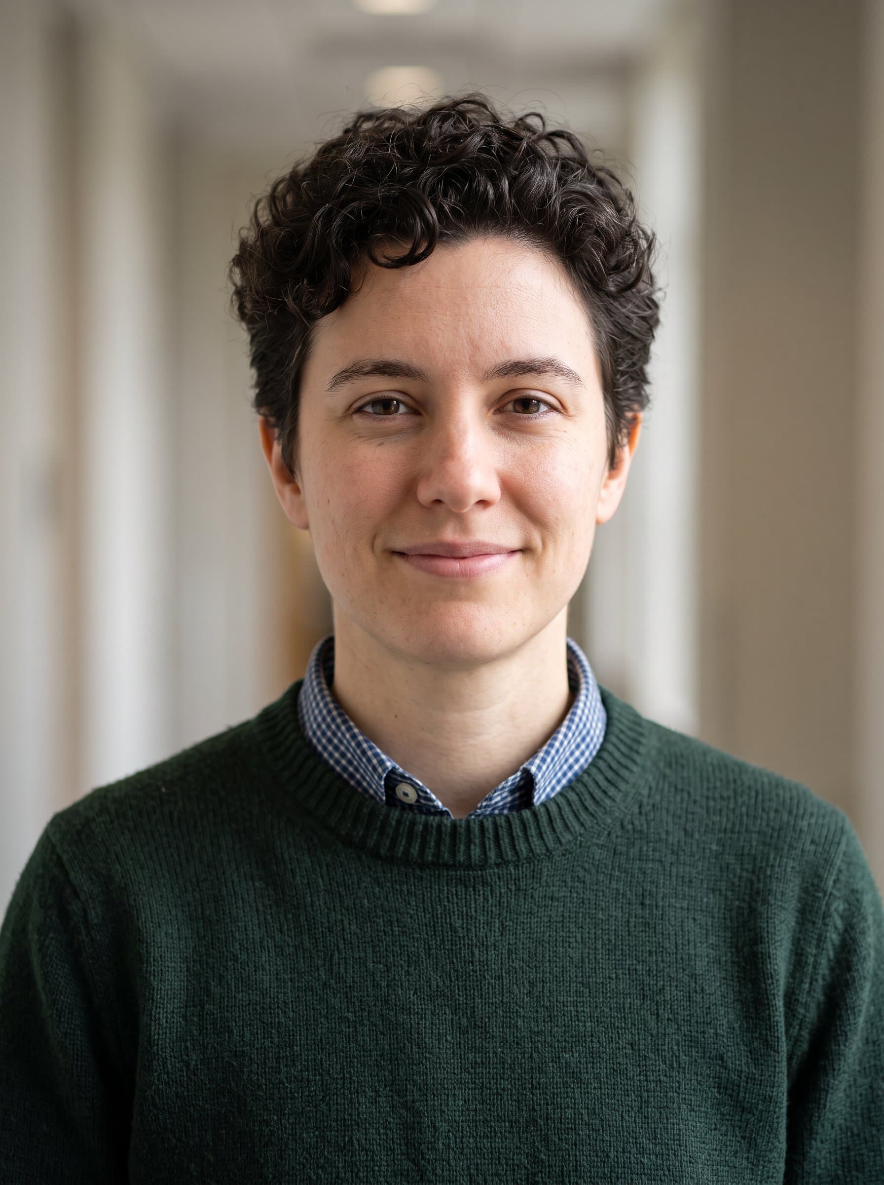 : Produce a faculty member headshot with a gender-neutral academic, in a dark green sweater over a collared shirt. Neutral background and gentle lighting for a polished university profile image.