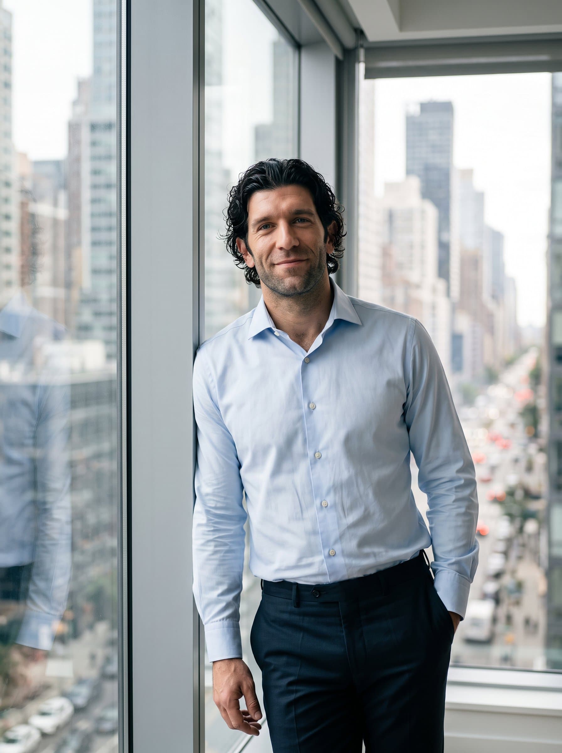 : A man in a formal dress shirt poses by a large window with city views, smiling gently. The background bokeh effect highlights their approachable and professional presence.