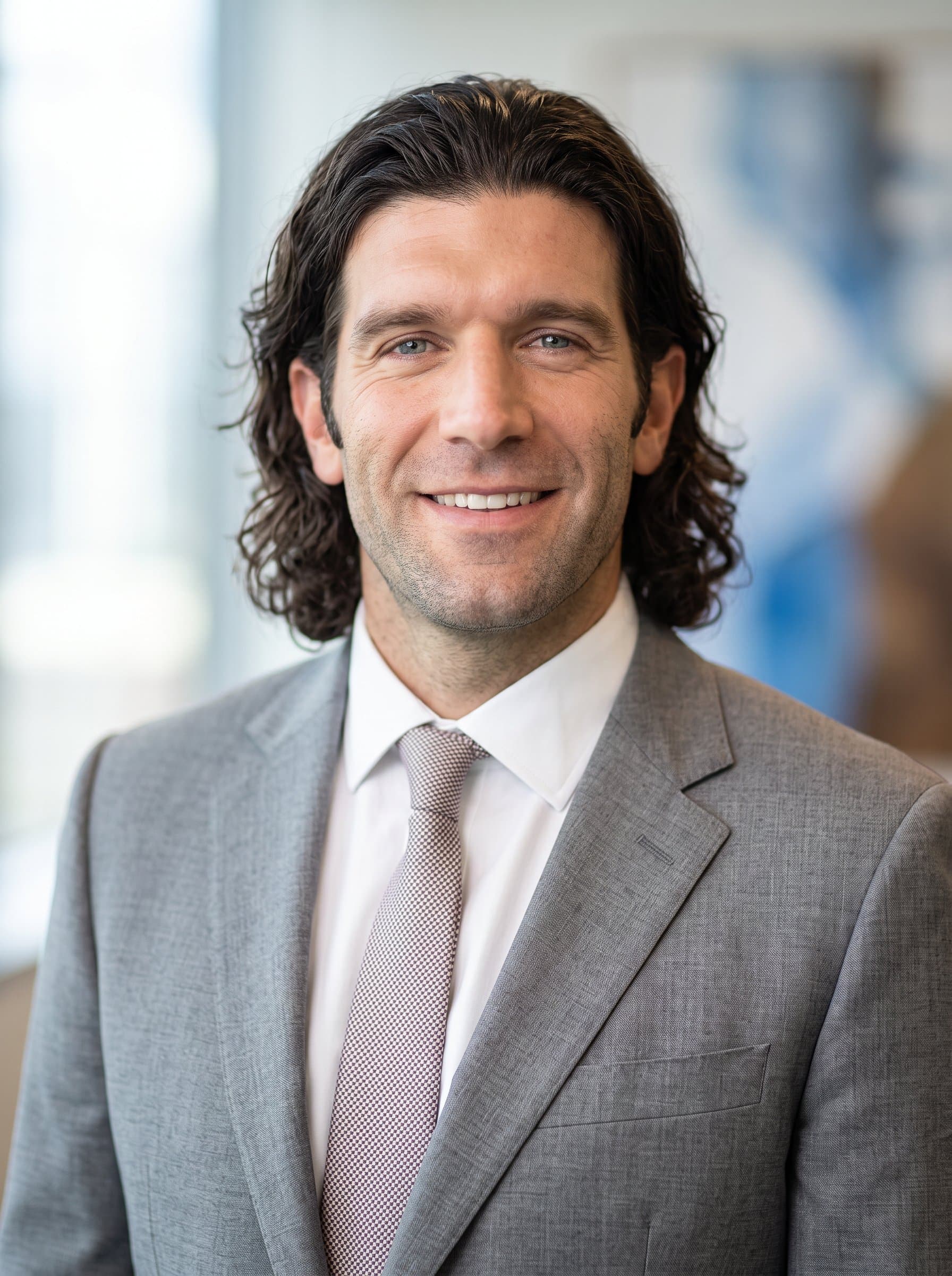 : Create a polished, corporate headshot with a soft focus. The man is wearing a light-colored suit, standing against a gentle, abstract background, exuding professionalism and approachability.