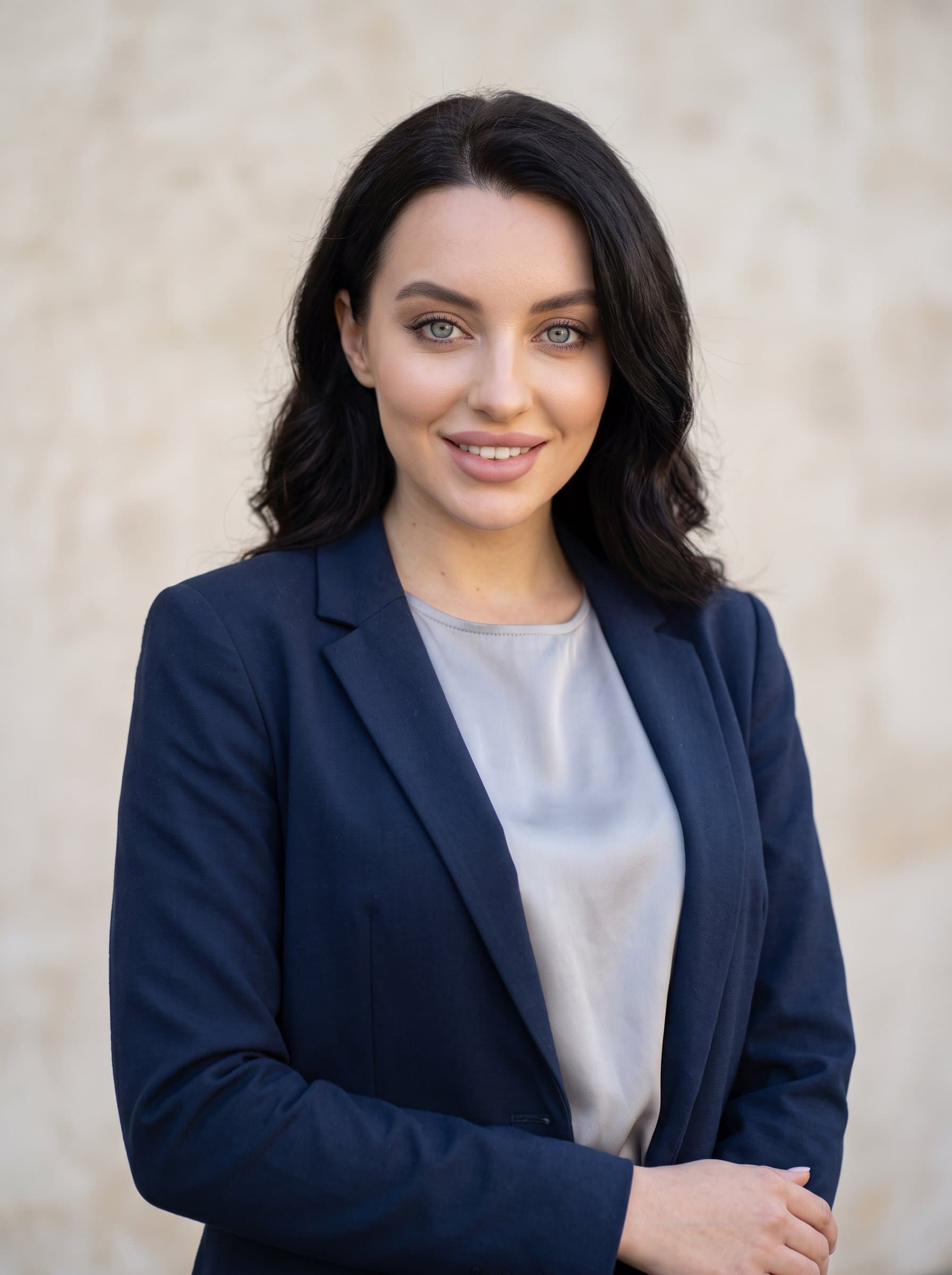 Portray a headshot featuring direct gaze and a relaxed posture. The woman stands against a subtly textured background, wearing a smart-casual outfit that reflects a balance of professionalism and approachability.
