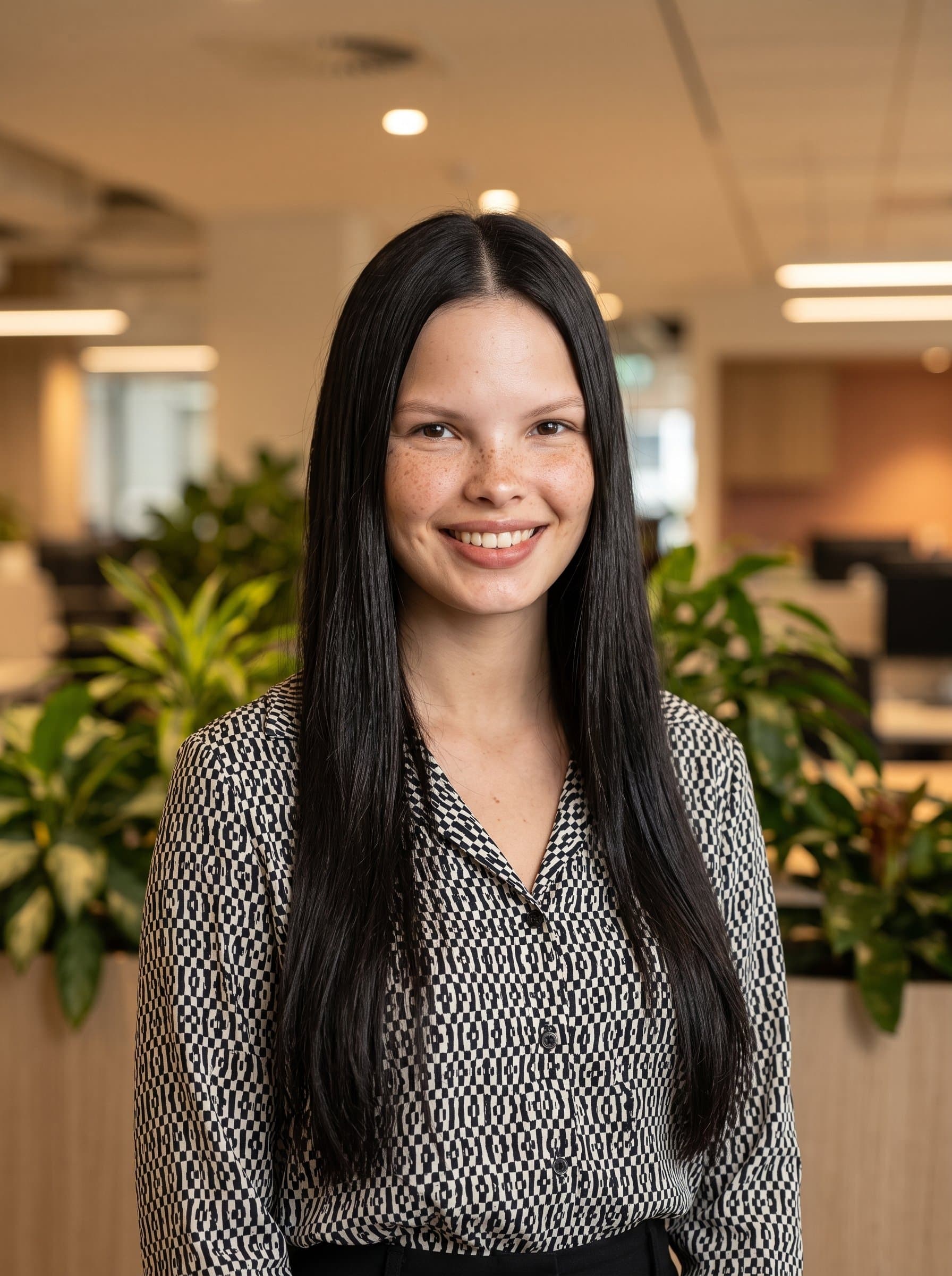 composed headshot of an woman in a monochrome shirt, standing in front of office plants. Warm lighting creates a positive atmosphere, enhancing the woman’s professionalism for use in corporate settings.