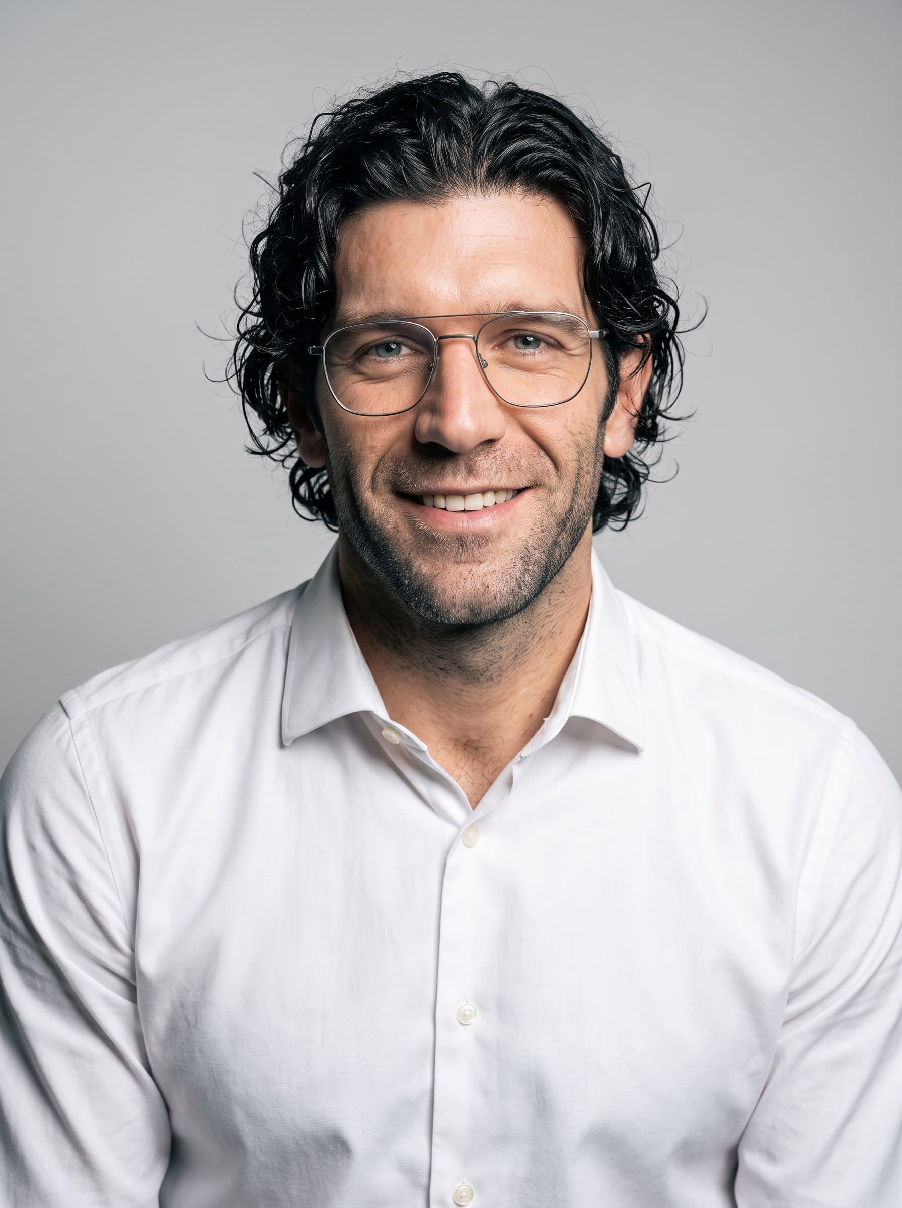 Generate an AI headshot for set in a studio with a pale grey background. The man, with medium-length hair and glasses, wears a crisp, white shirt, exuding warmth and professionalism.