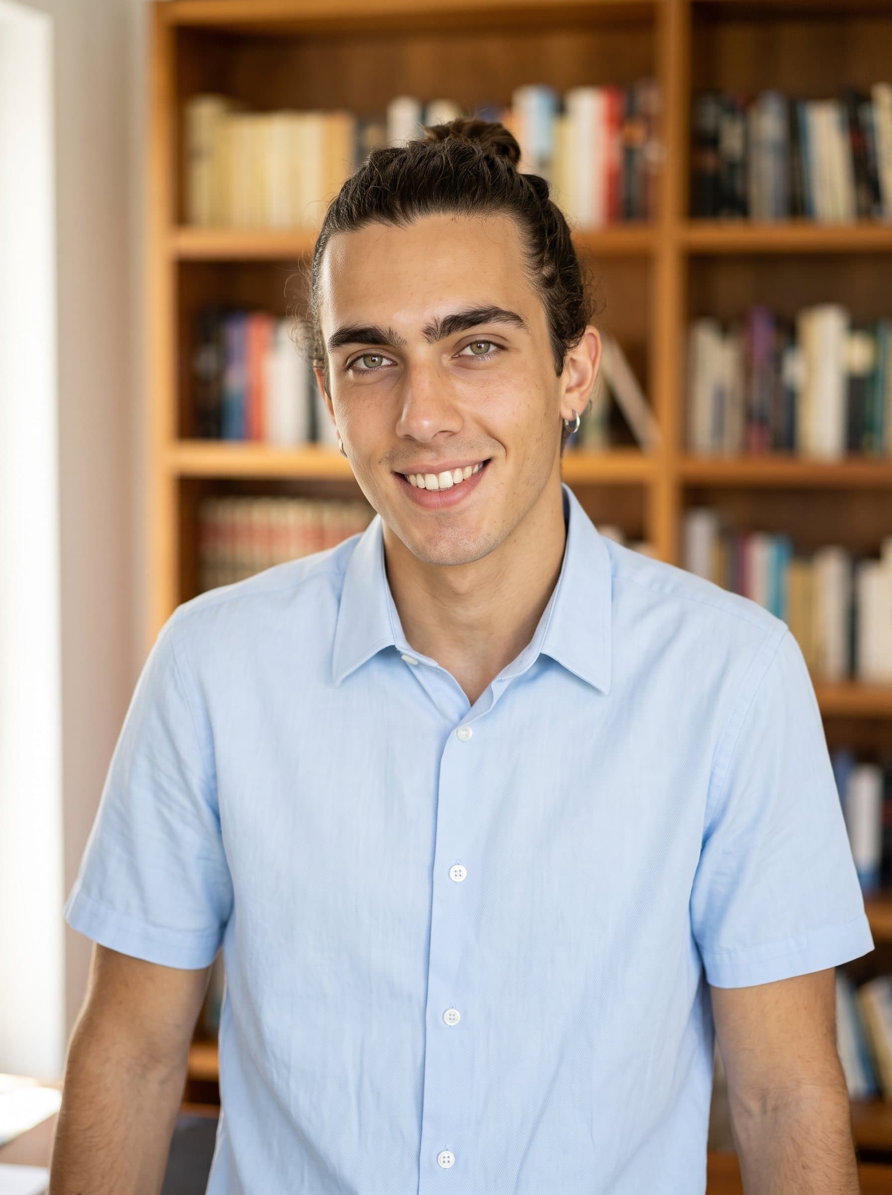 Craft a high-resolution image of with a neat bun hairstyle and a pastel collared shirt. The man stands before a softly blurred bookshelf, providing a warm and intelligent aura.