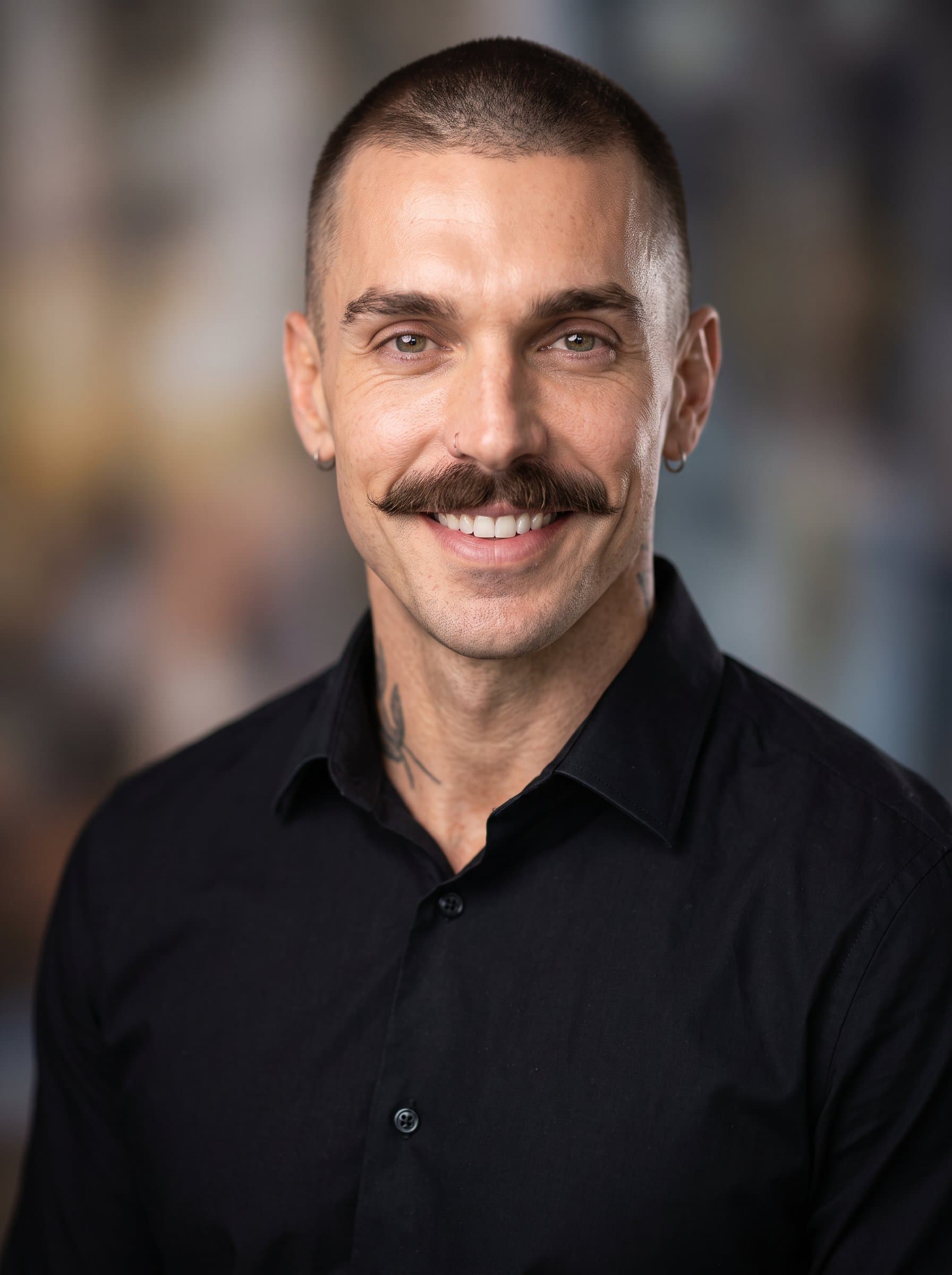 Create a professional headshot of featuring an man with a buzz cut, wearing a formal black shirt. The background is a subtle, abstract blur to ensure focus on the man's welcoming expression.