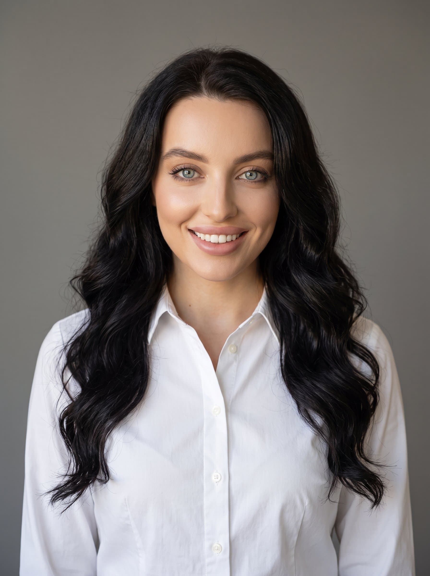 : Capture a classic professional headshot with a neutral gray background. The woman is centered, confidently smiling, wearing a crisp white shirt. Lighting is natural, emphasizing a warm, approachable demeanor for a Twitter profile.