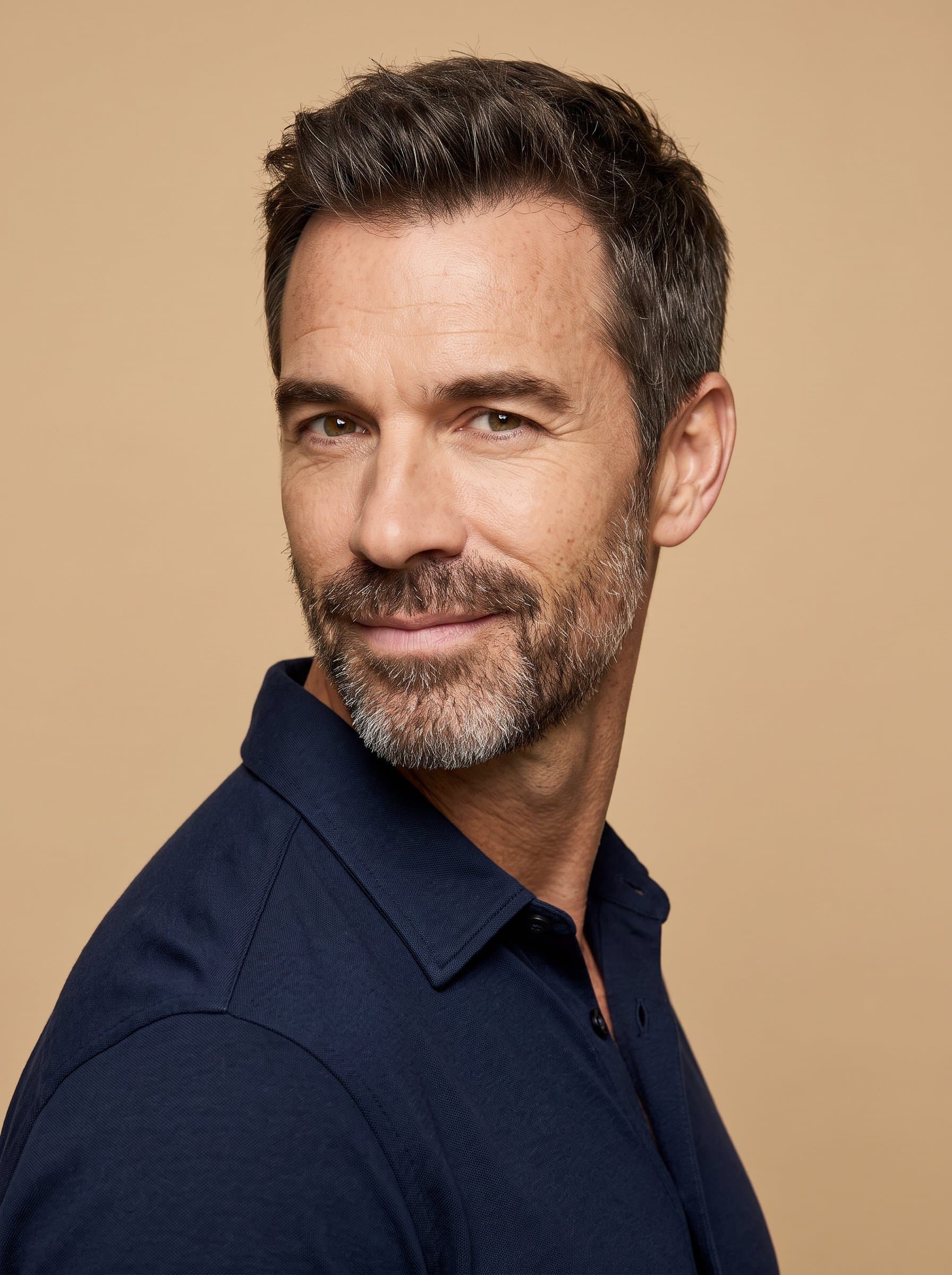 Compose a image of a man subtly smiling, facing slightly sideways. The unobtrusive sand-colored background elevates their facial clarity, making their engaging demeanor ideal for digital use.