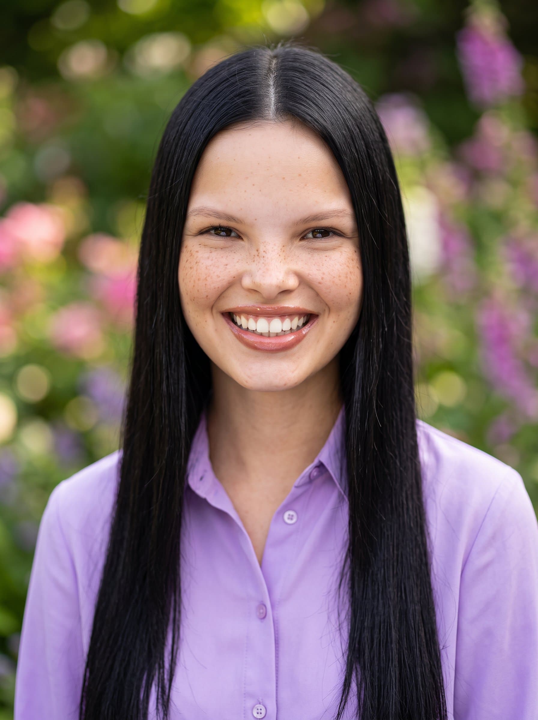 Design a headshot of a woman with an expressive smile, wearing a lavender button-up shirt. A blurred garden setting in the background brings a touch of nature to the professional portrayal.