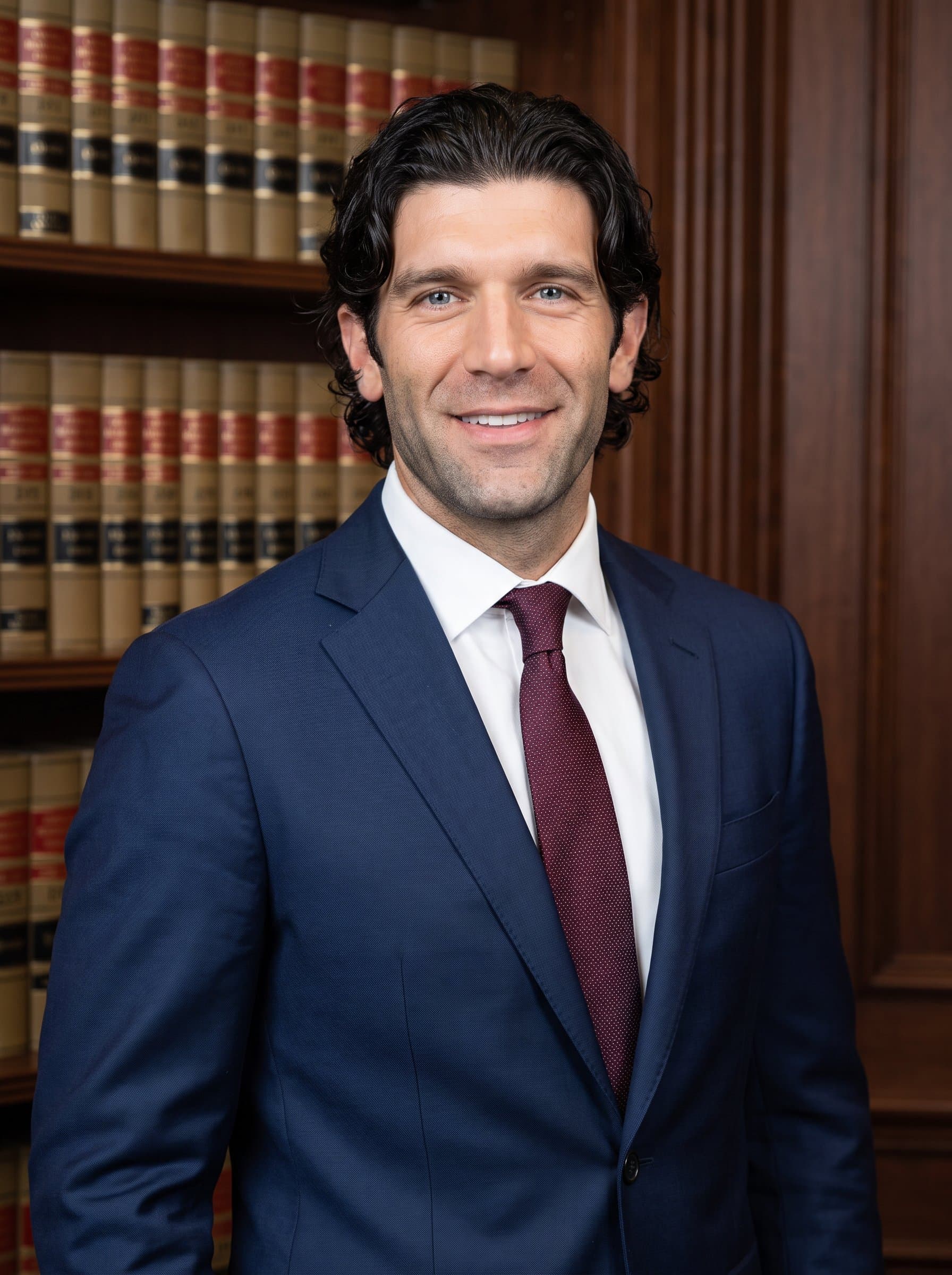 : Legal professional headshot, featuring an authoritative man in formal business attire, with a law library background, exuding confidence and competence for a Glassdoor profile.